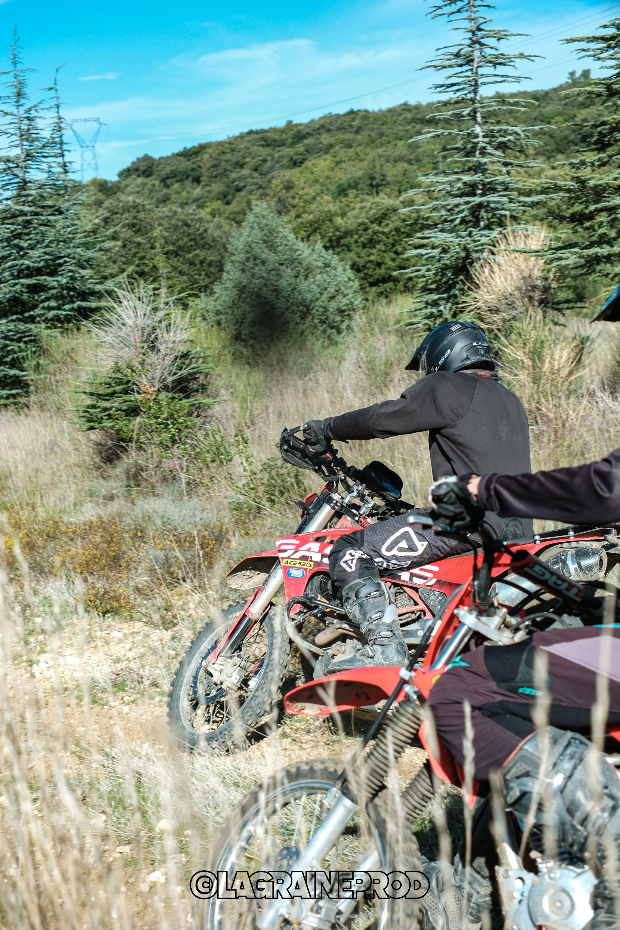 Deux motocyclistes en tenue et casque roulant à travers un paysage boisé avec des conifères et des collines en arrière-plan.