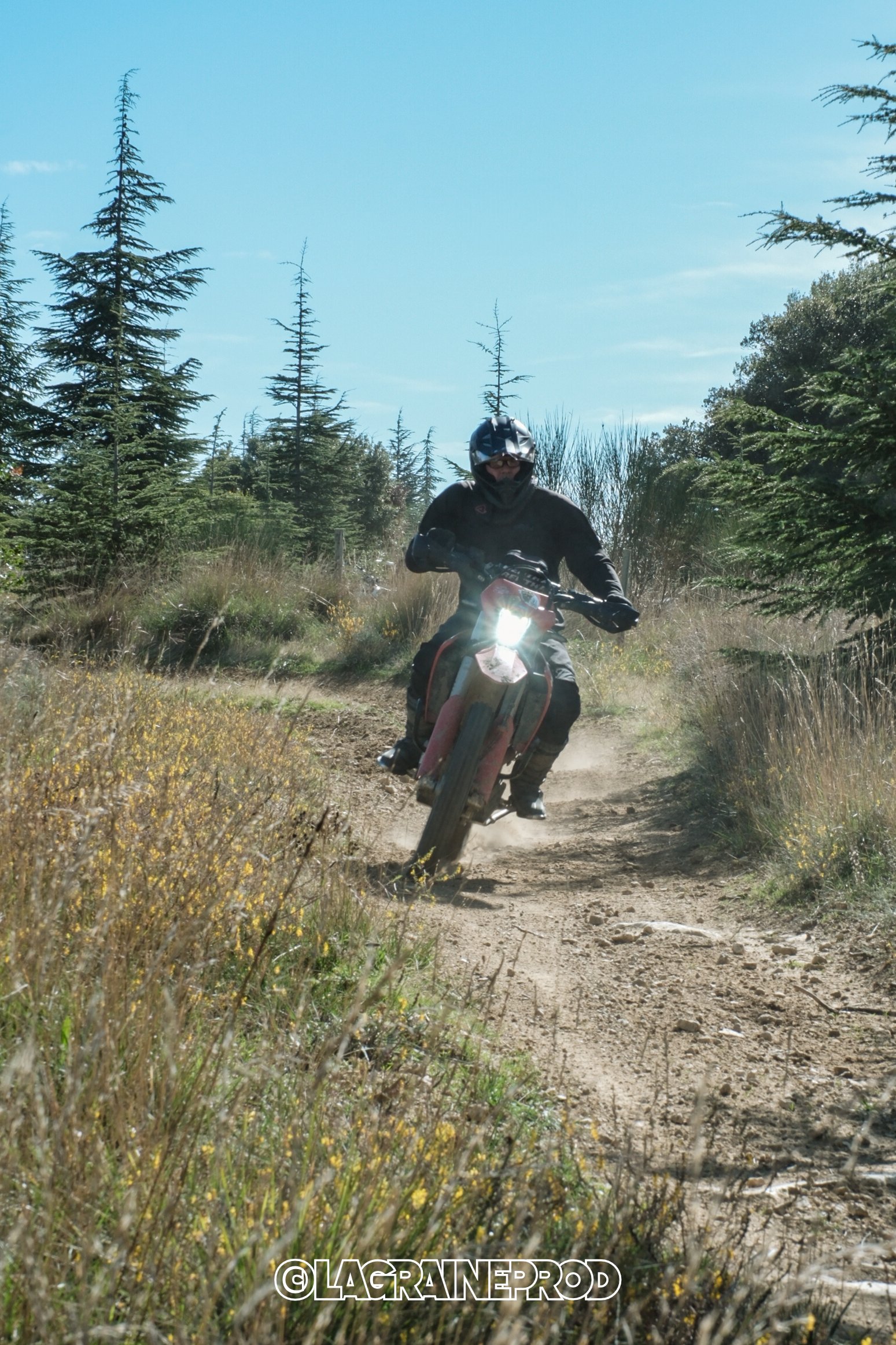 Un motocycliste portant un casque noir et des vêtements noirs, roulant sur un sentier de terre au milieu de la forêt avec des arbres verts, sous un ciel bleu.