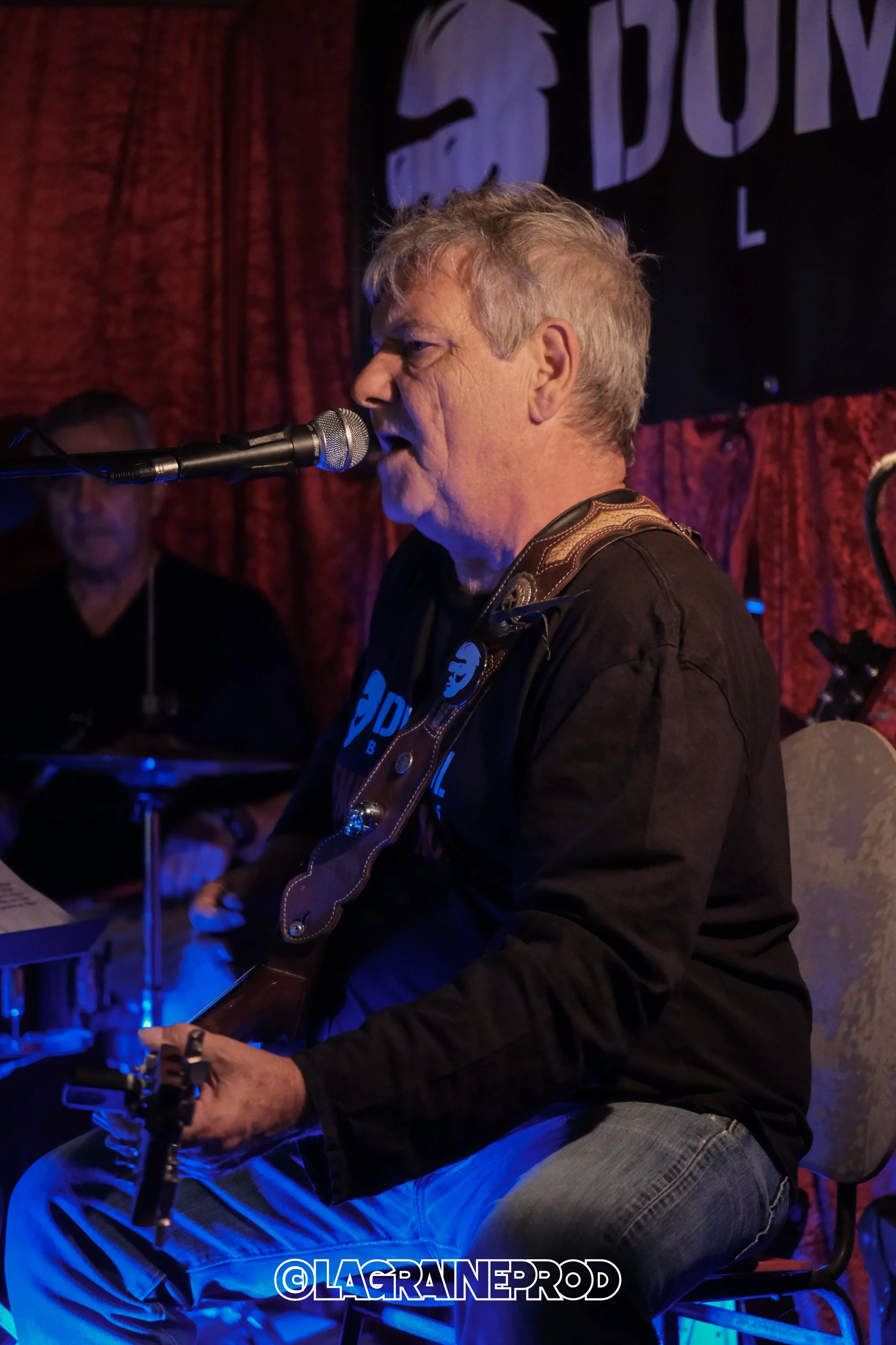Un homme joue de la guitare et chante dans un club de musique, avec un microphone devant lui et un batteur en arrière-plan, ambiance tamisée et scène avec rideaux rouges.