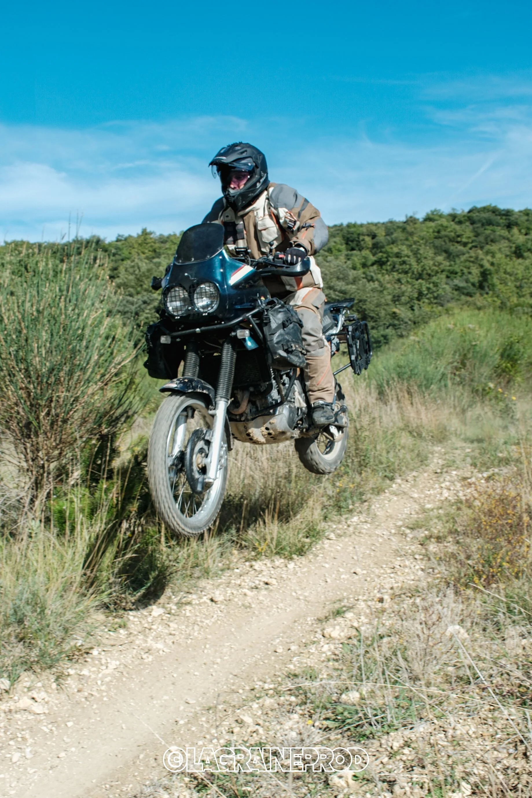 Un motocycliste portant un casque et un équipement de protection saute un obstacle sur un chemin en dirt, avec des collines verdoyantes en arrière-plan sous un ciel bleu.