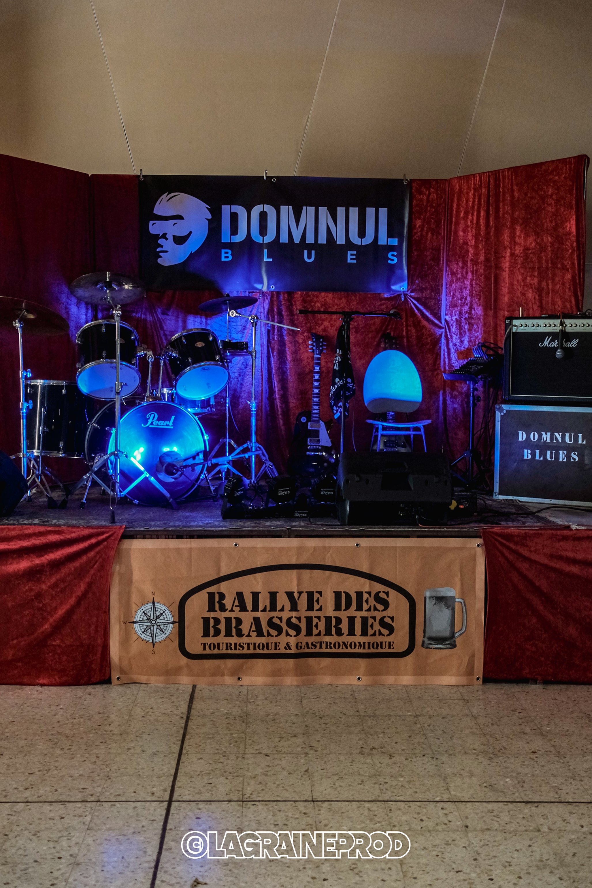Une scène de musique avec une batterie, une guitare, un clavier, une chaise et un ampli, sur une scène avec un rideau rouge. Il y a une bannière "DOMNUL. BLUES" en arrière-plan et un panneau "RALLYE DES BRASSERIES" en devanture.
