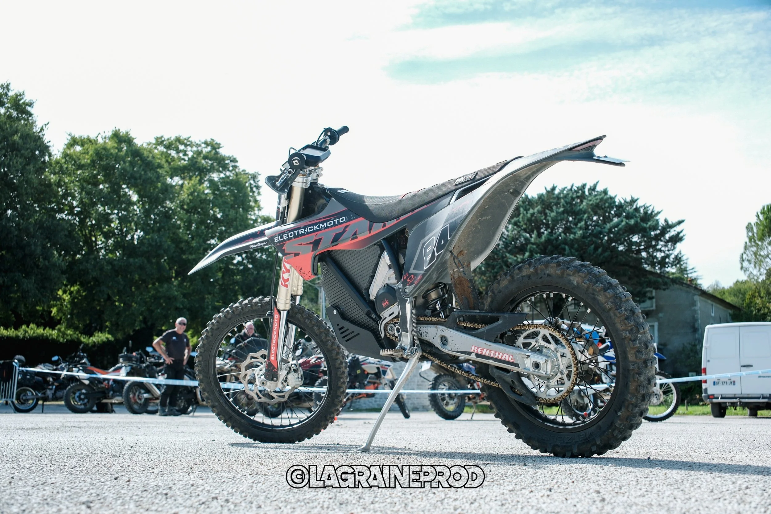 Une moto électrique enduro avec un cadre noir, des pneus tout-terrain, et des logos 'Electrickmoto' et 'Renthal' visible, stationnée sur le sol avec des personnes et d'autres motos en arrière-plan.