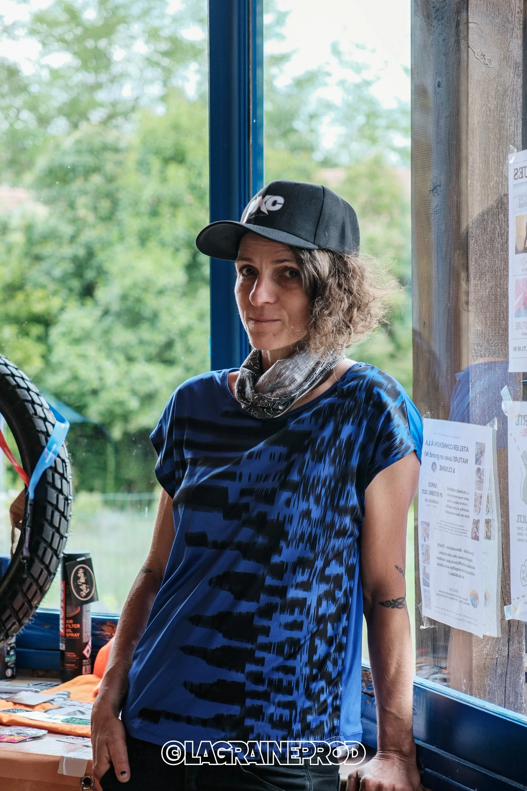 Une femme avec des cheveux bouclés, portant une casquette noire, un bandana gris et un t-shirt bleu et noir, se tient à côté d'une fenêtre. Elle pose près d'une table à l'intérieur d'un lieu en bois, avec des affiches collées au mur.