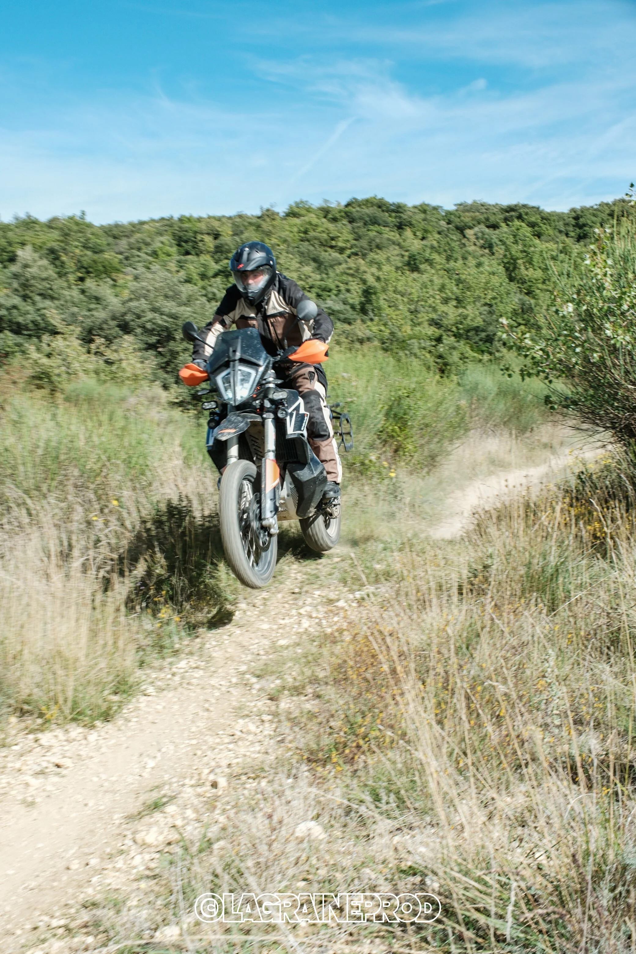 Un motard portant un casque noir et des vêtements de protection, chevauchant une moto tout-terrain sur un chemin de terre en pleine nature avec des montagnes verdoyantes en arrière-plan.