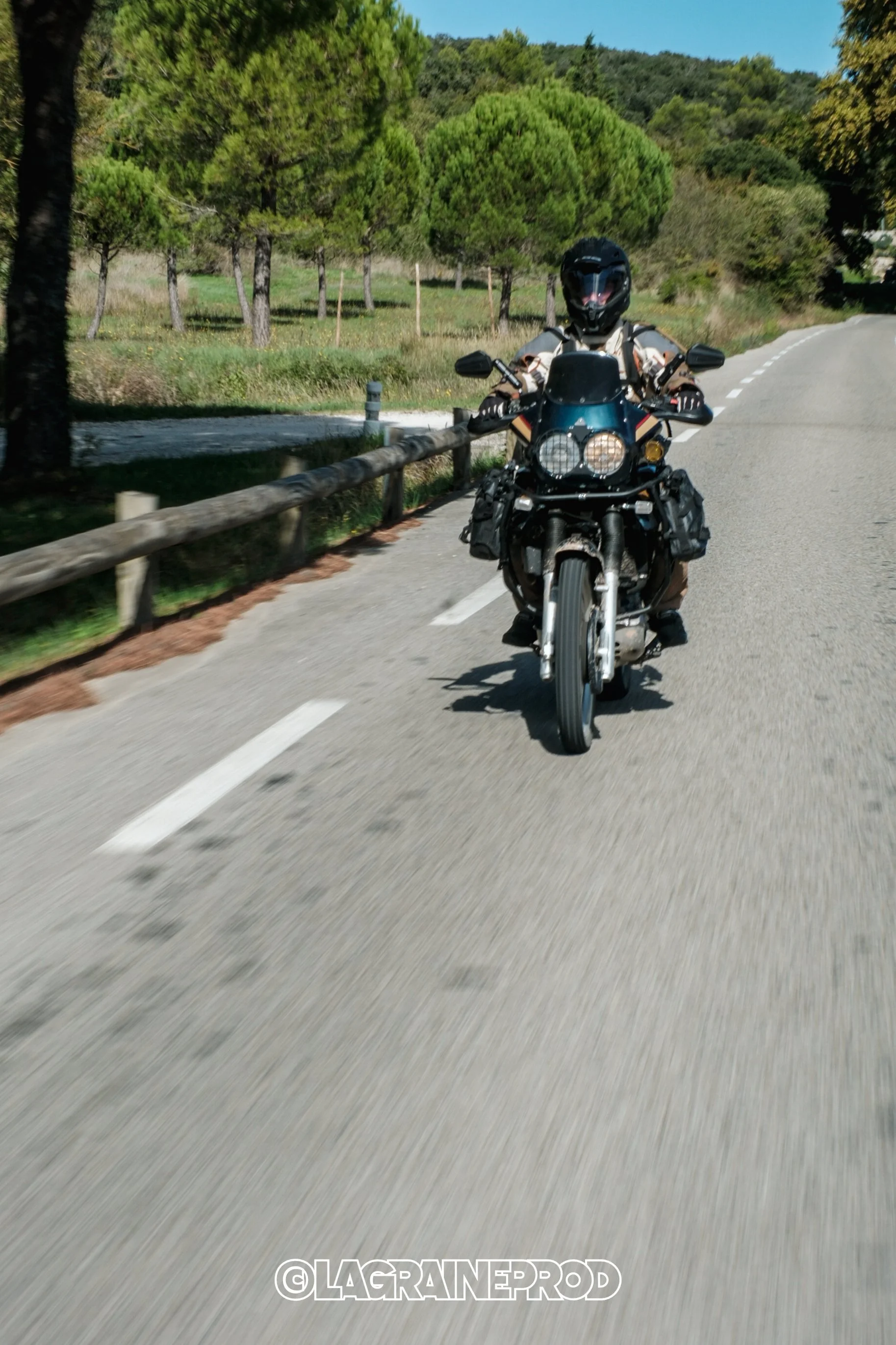 Motard portant un casque noir, roulant sur une route bordée d'arbres verts, avec un ciel bleu.