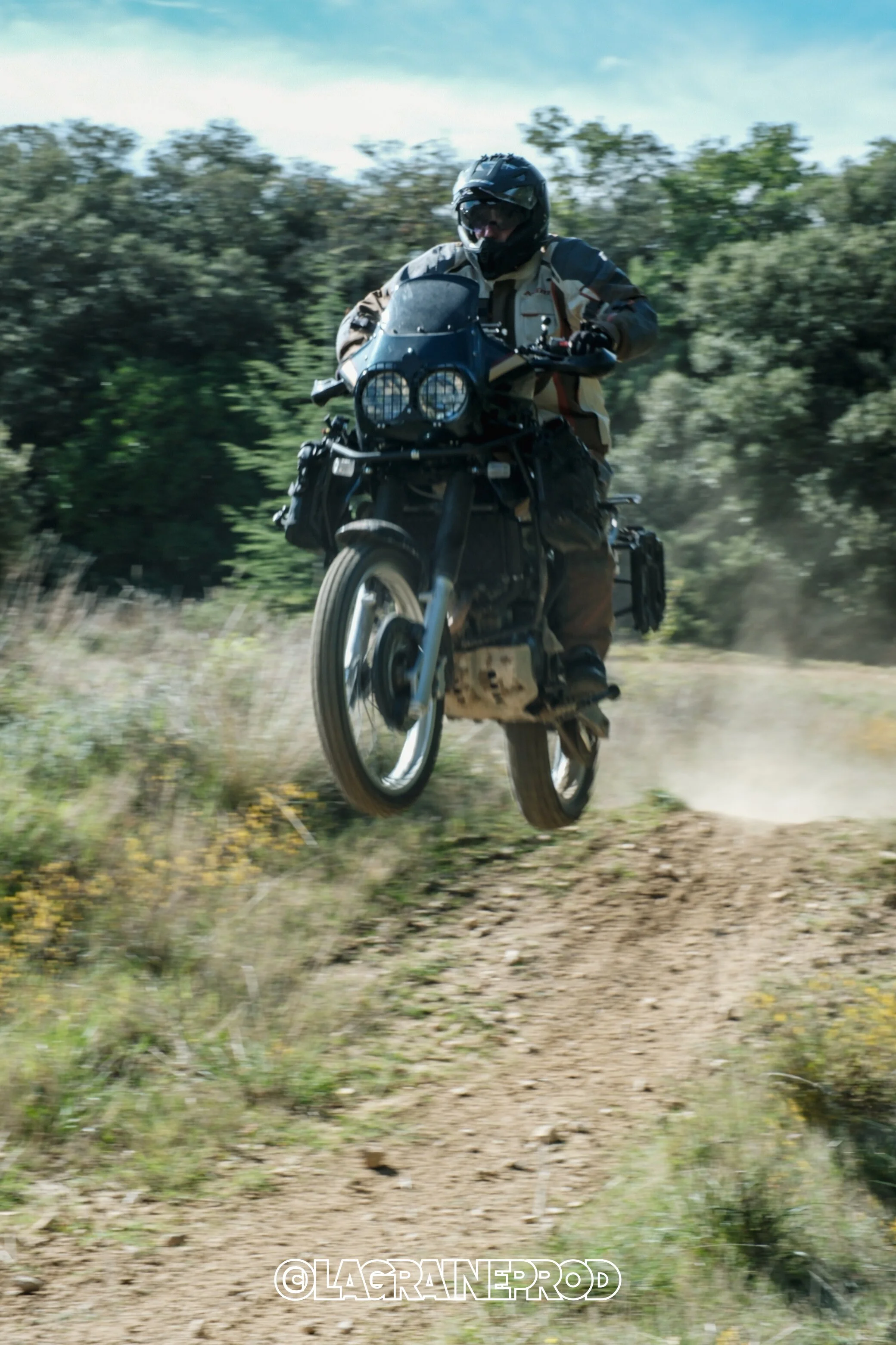 Un motard portant un casque et une veste de protection, sautant avec sa moto tout-terrain sur un chemin de terre dans un environnement forestier.