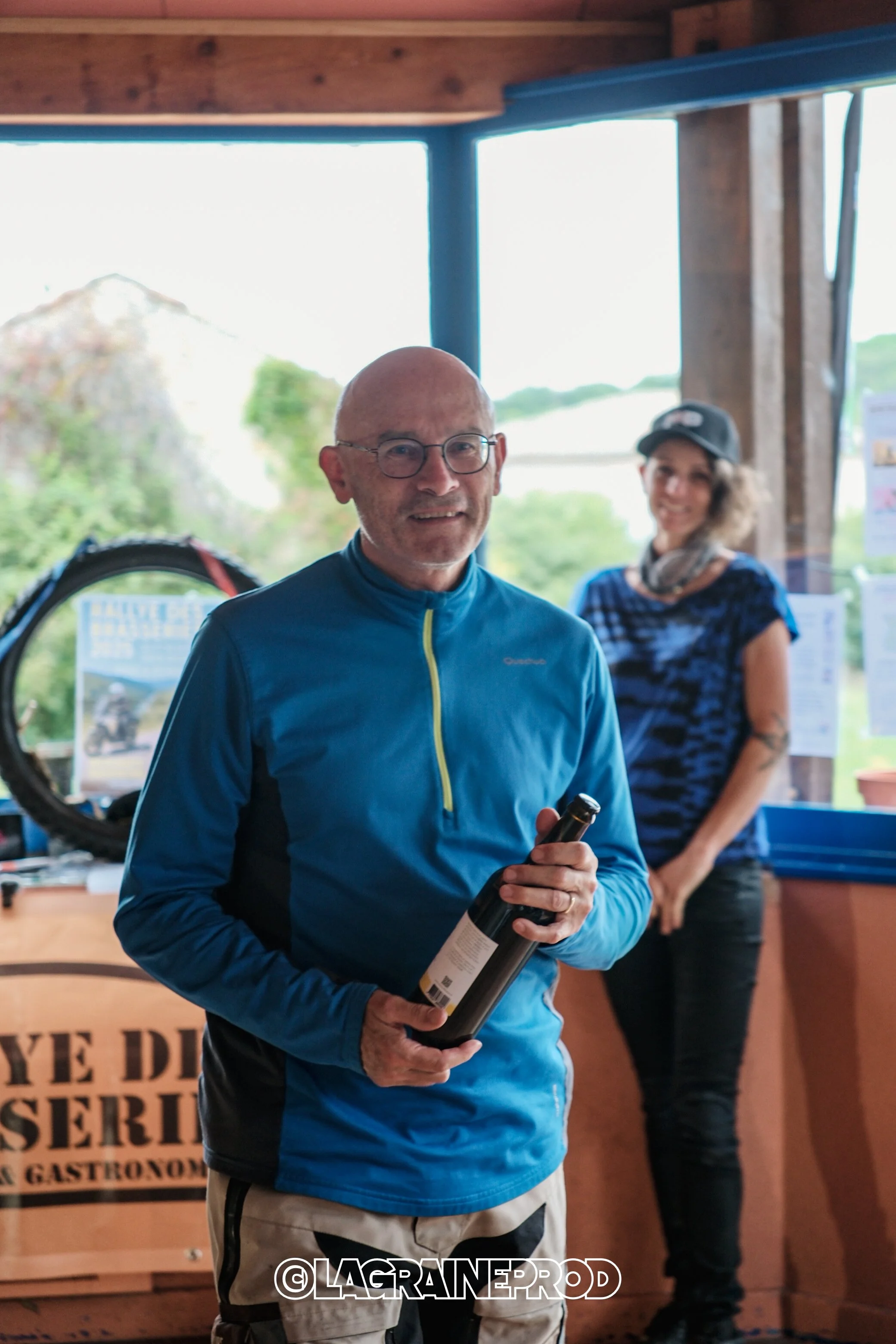 Un homme tenant une bouteille de vin dans un environnement intérieur, avec une femme en arrière-plan, tous deux souriants.