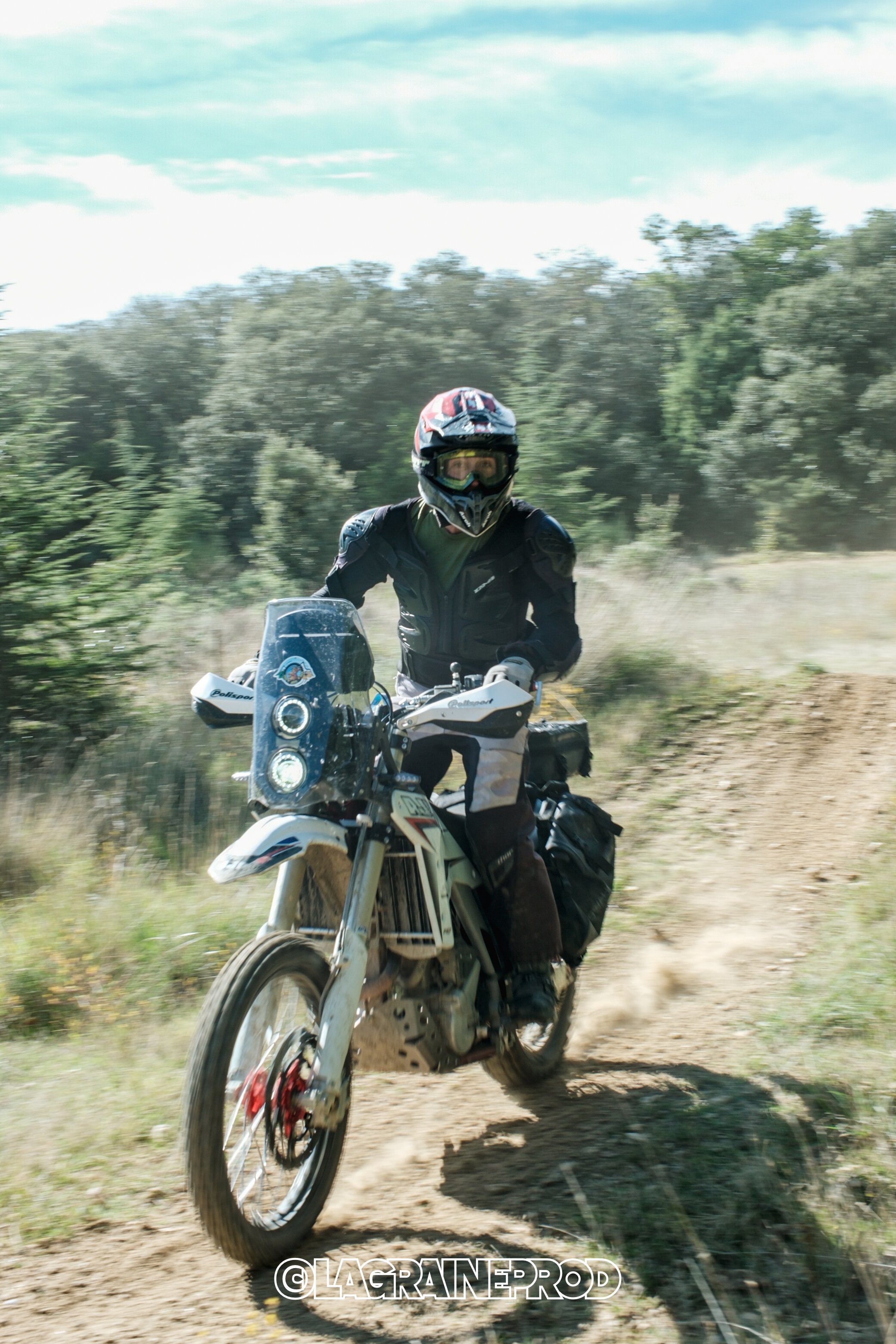 Motard en équipement de protection, portant un casque et des vêtements de moto, roulant sur un chemin de terre dans la nature avec des arbres en arrière-plan.