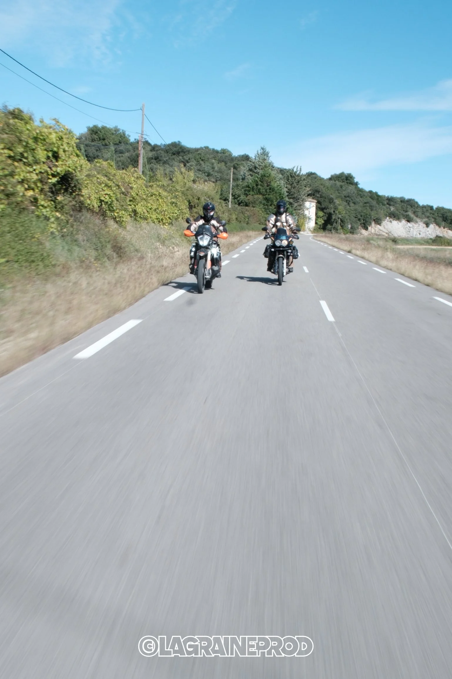 Deux motards portant un équipement de protection roulent sur une route de campagne bordée d'arbustes et de collines, sous un ciel bleu clair.