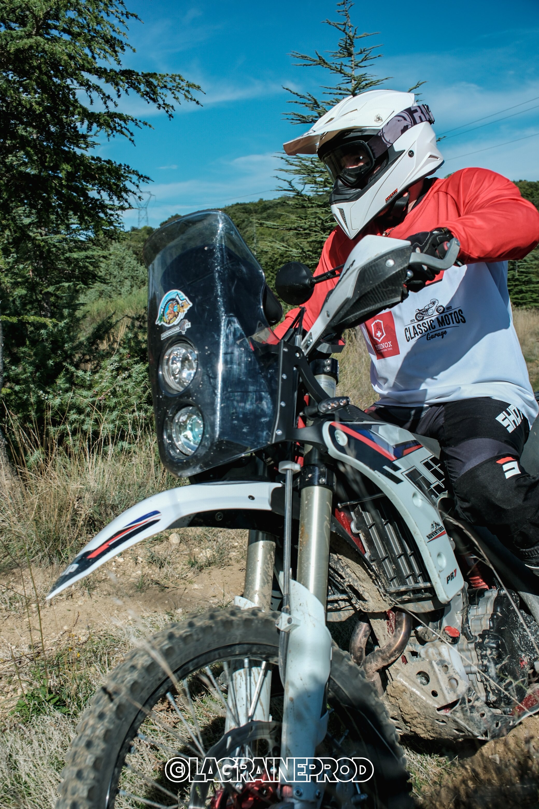 Un motard portant un casque blanc, un maillot blanc avec des logos et un pantalon noir, en train de rouler dans un environnement naturel avec des arbres et un ciel bleu.