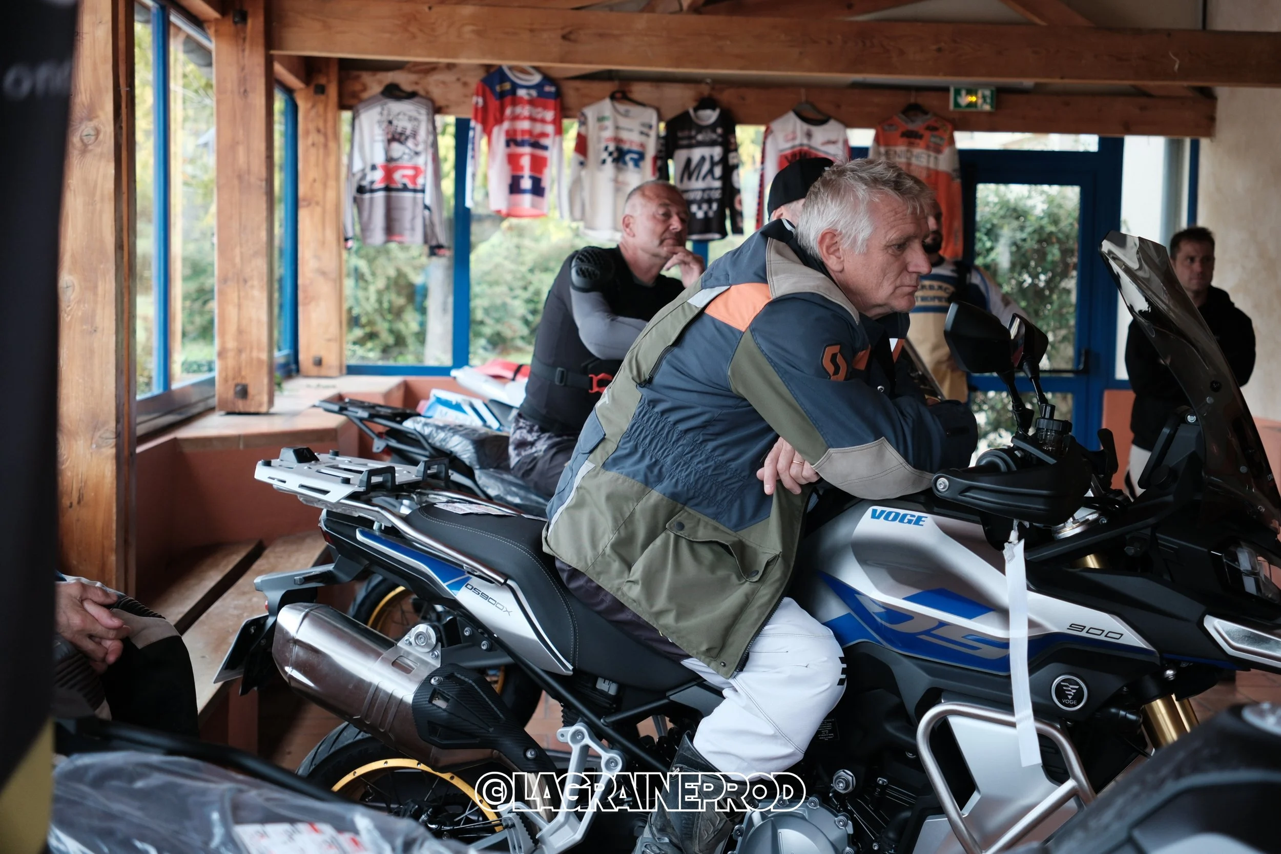 Plusieurs personnes dans un atelier de moto, avec un homme assis sur une moto Voge 900, portant une veste colorée, à l'intérieur d'une pièce en bois. Des maillots de course sont suspendus au mur.