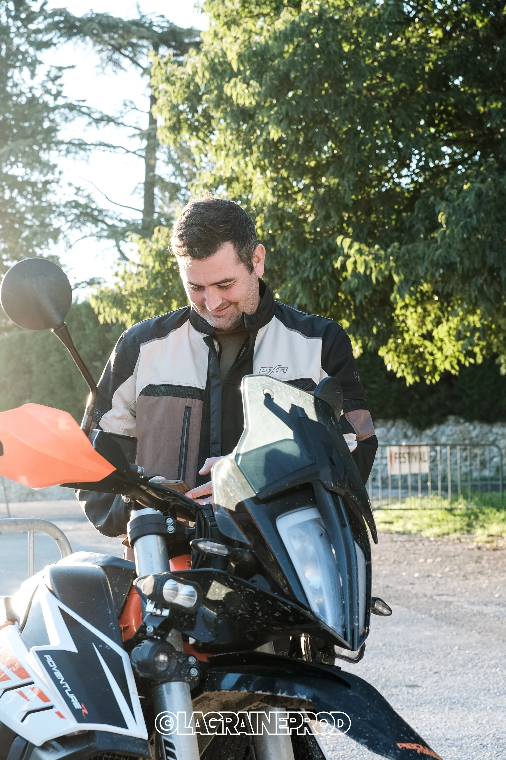 Un homme souriant portant une veste de moto, debout à côté d'une moto noire avec des accents orange, en plein air avec des arbres en arrière-plan.