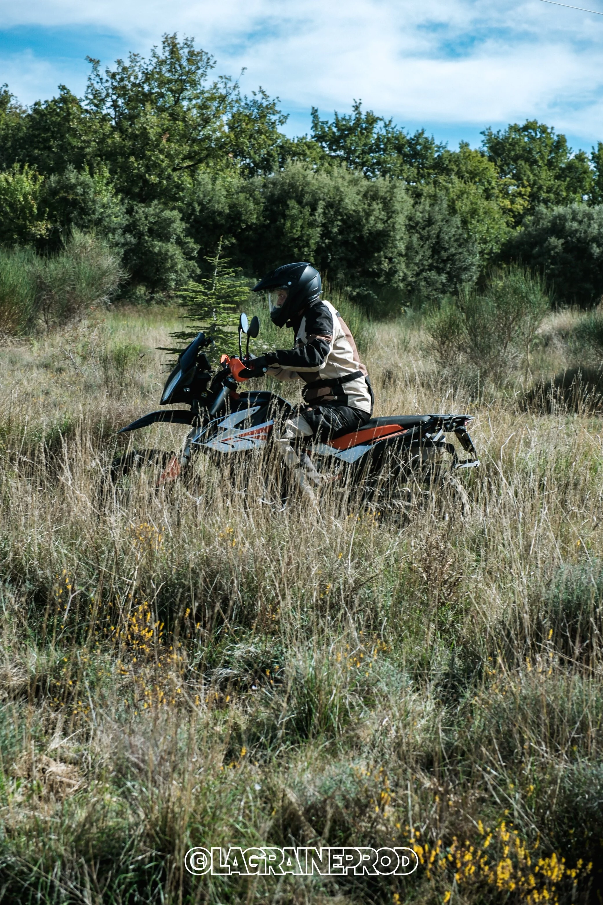 Une personne portant un casque et une veste en équilibre sur une moto tout-terrain dans un champ herbeux avec des arbres en arrière-plan.