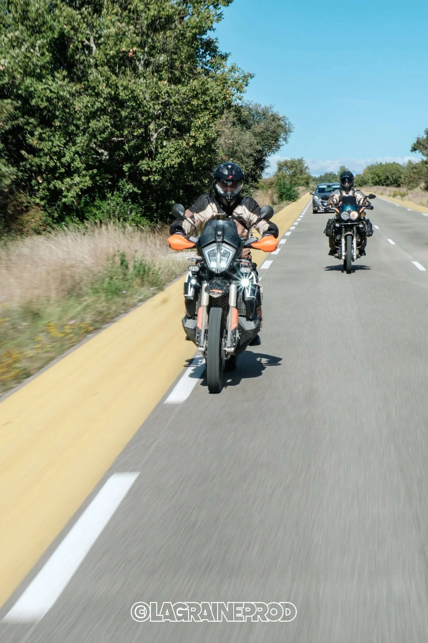 Deux motocyclistes portant des casques et des vestes, roulant sur une route de campagne sous un ciel bleu, avec une voiture en arrière-plan.