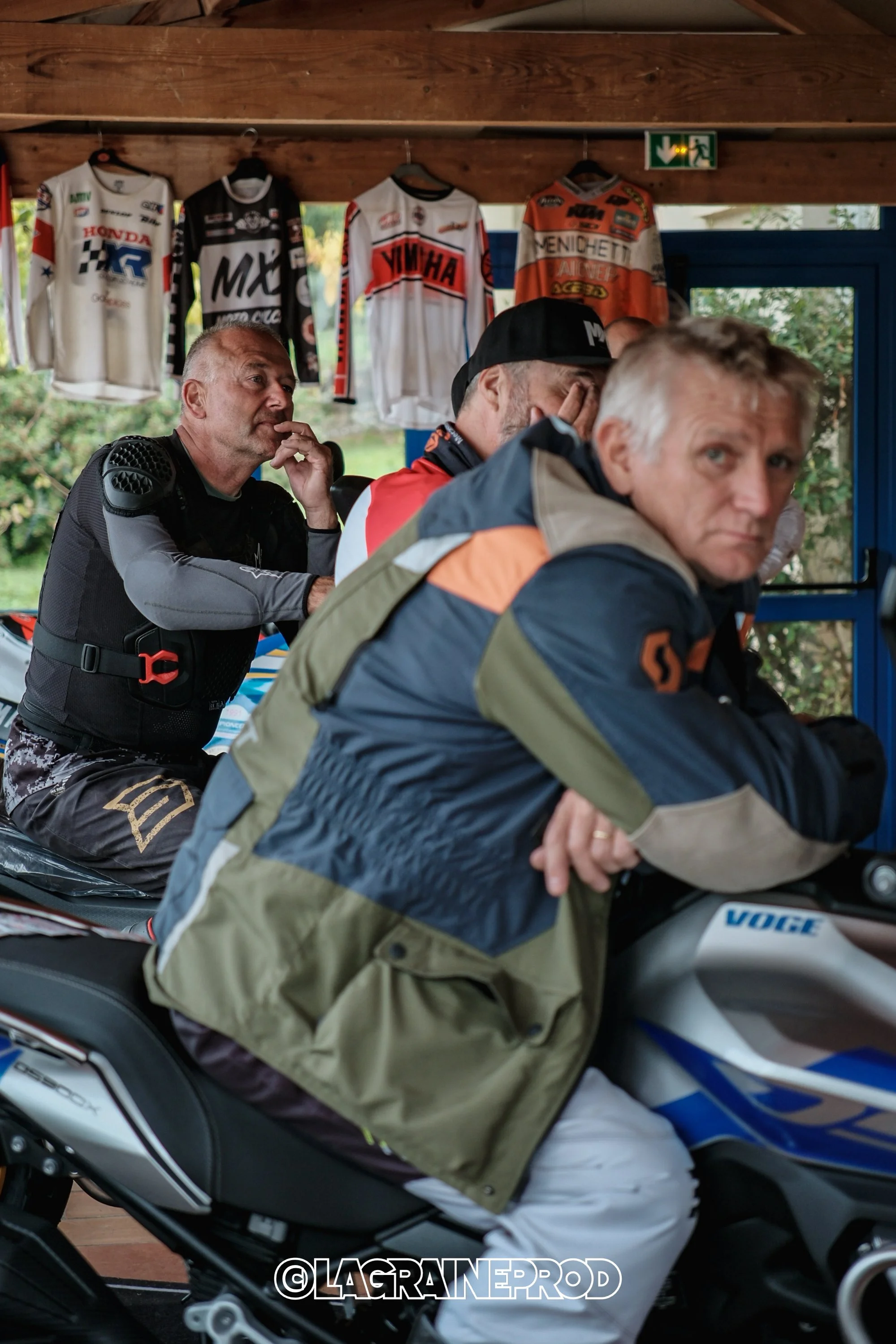 Trois hommes assis sur une moto, avec des vêtements de moto, dans un espace intérieur avec des maillots de motocyclisme suspendus au mur en arrière-plan.