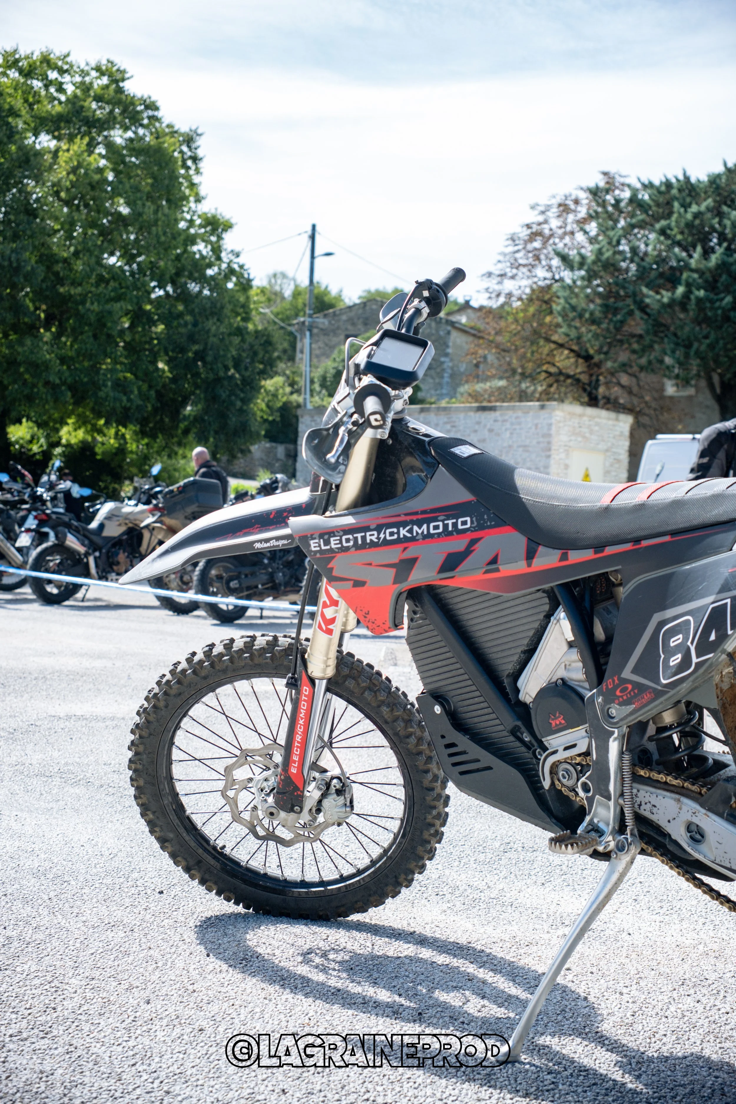Moto électrique tout-terrain noire avec des accents rouges et des pneus à crampons, stationnée dans une aire extérieure avec d'autres motos en arrière-plan.