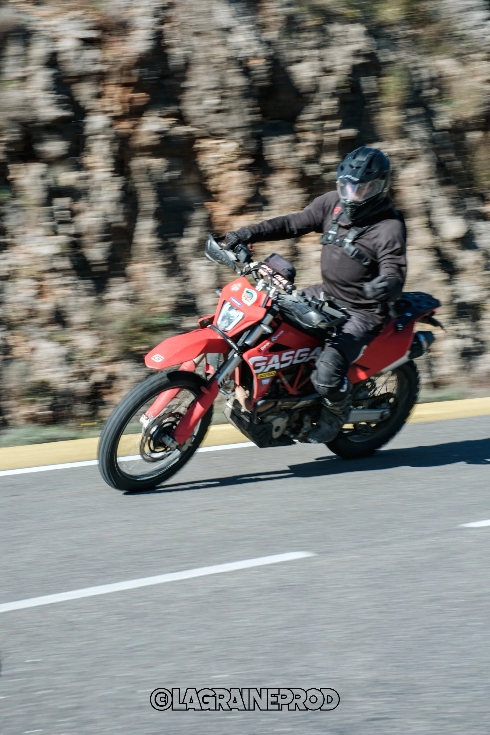 Un motard en tenue noire chevauche une moto rouge sur une route avec un rocher en arrière-plan.