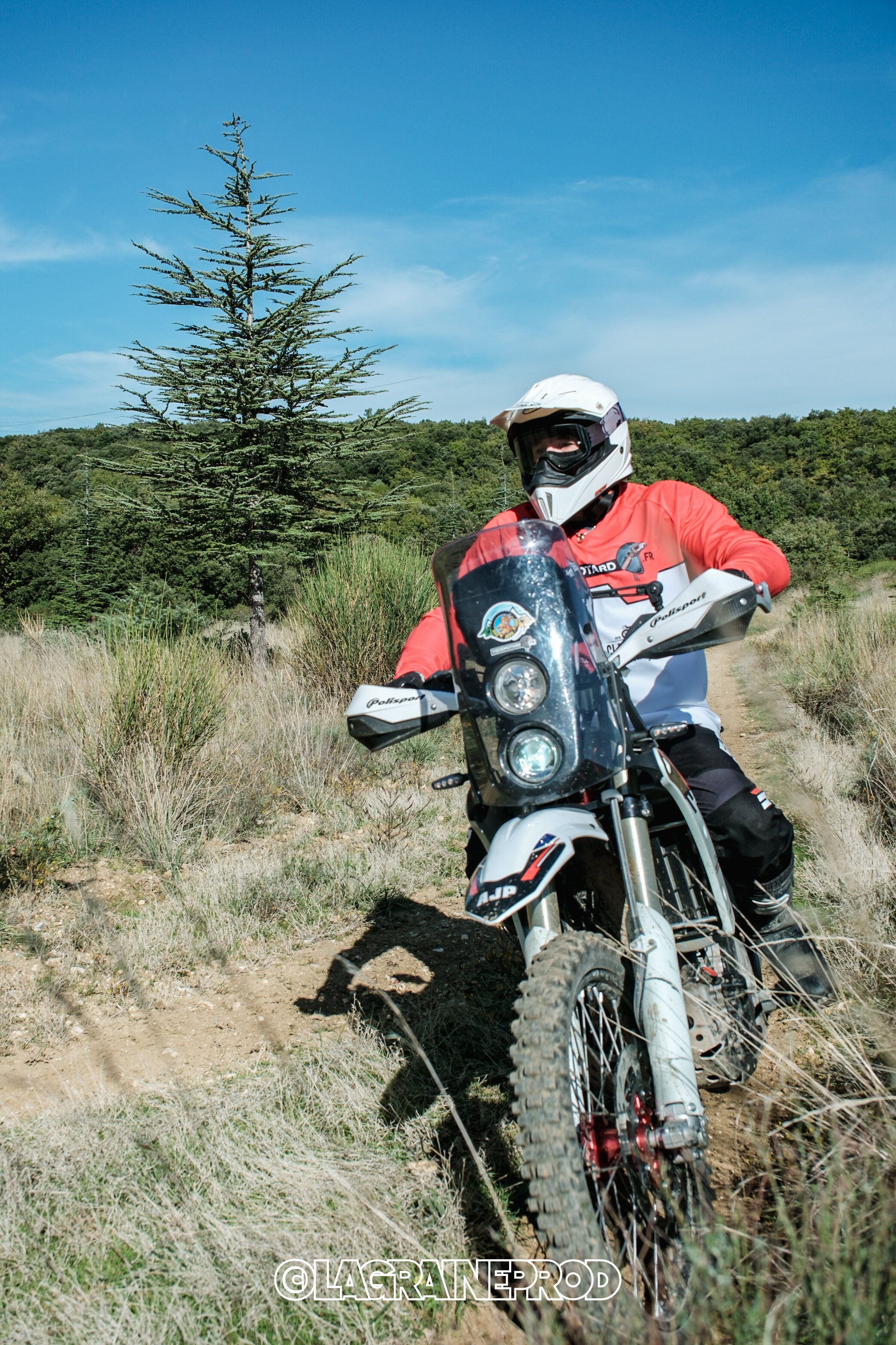 Personne portant un casque de moto et une veste rouge, conduisant une moto tout-terrain sur un sentier en plein air, avec des arbres et un ciel bleu.