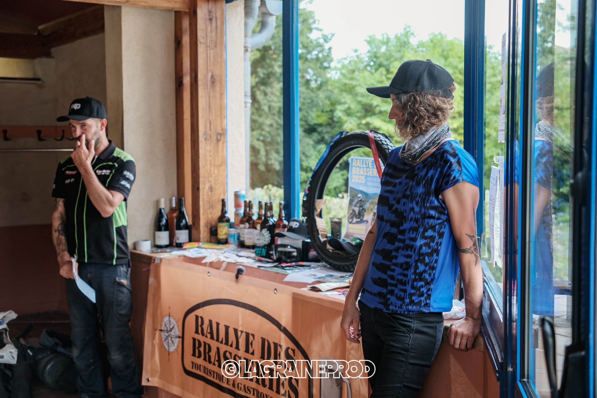 Une femme et un homme dans un espace intérieur, probablement une station ou un stand d'événement. La femme porte une casquette noire, un T-shirt bleu avec un motif noir, et pose en regardant dehors, tandis que l'homme en chemise noire et verte semble