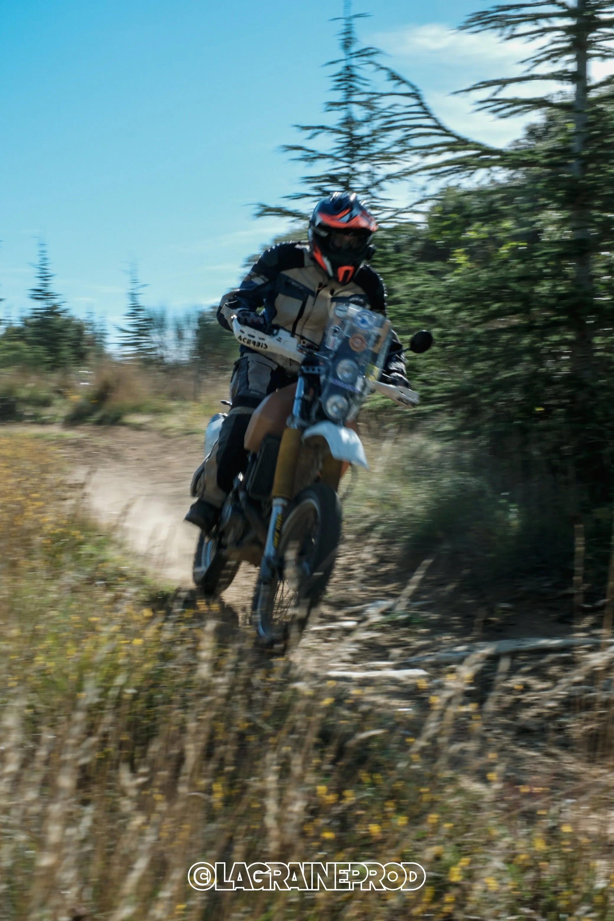 Un motard portant un casque noir et une tenue de moto tout-terrain fait du trail en forêt, la poussière s'élève derrière la roue.