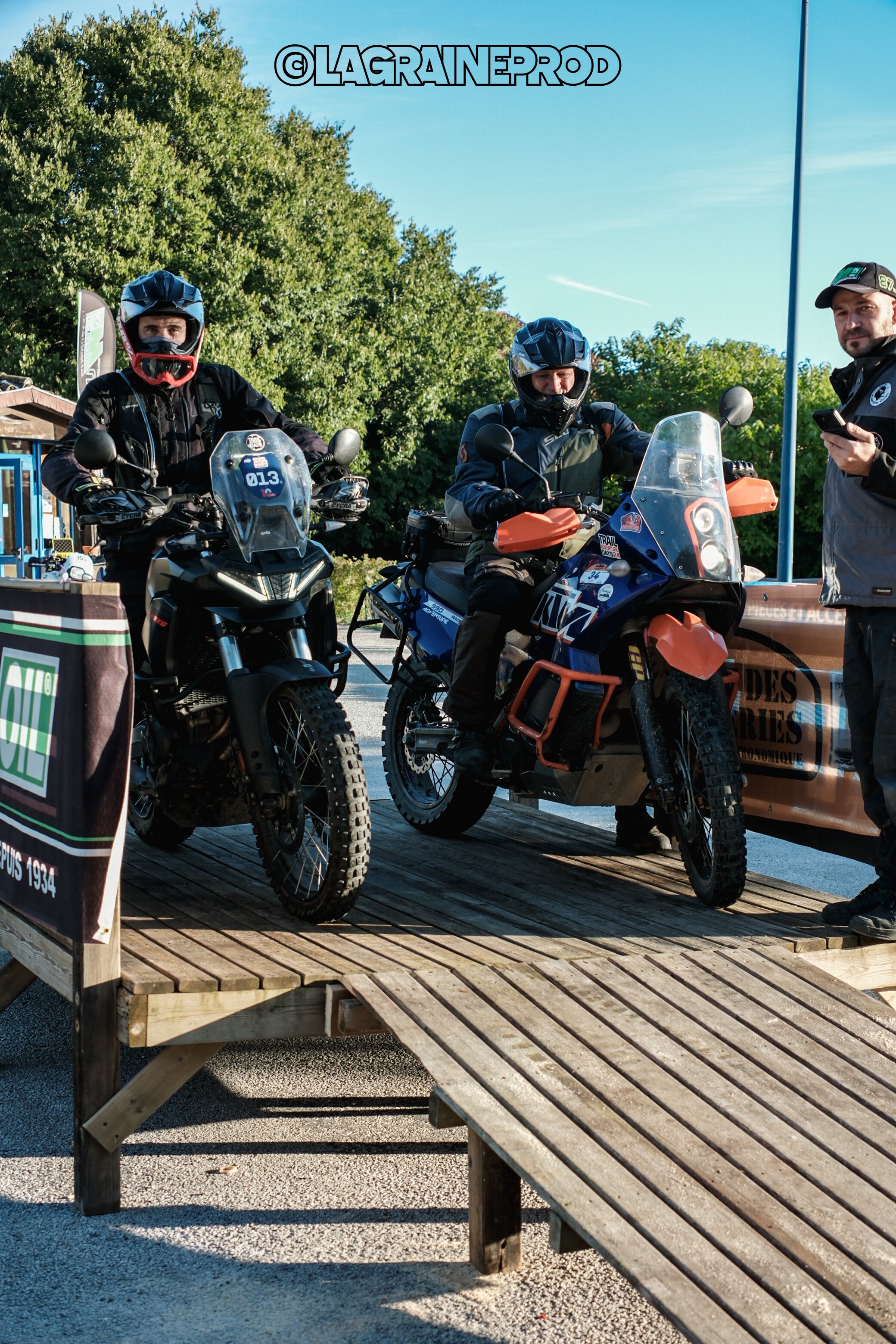 Deux motards en équipement de protection sont assis sur leurs motos tout-terrain, avec une personne debout à côté d'eux. La scène semble se dérouler lors d'un événement ou d'une compétition de moto, en plein air.