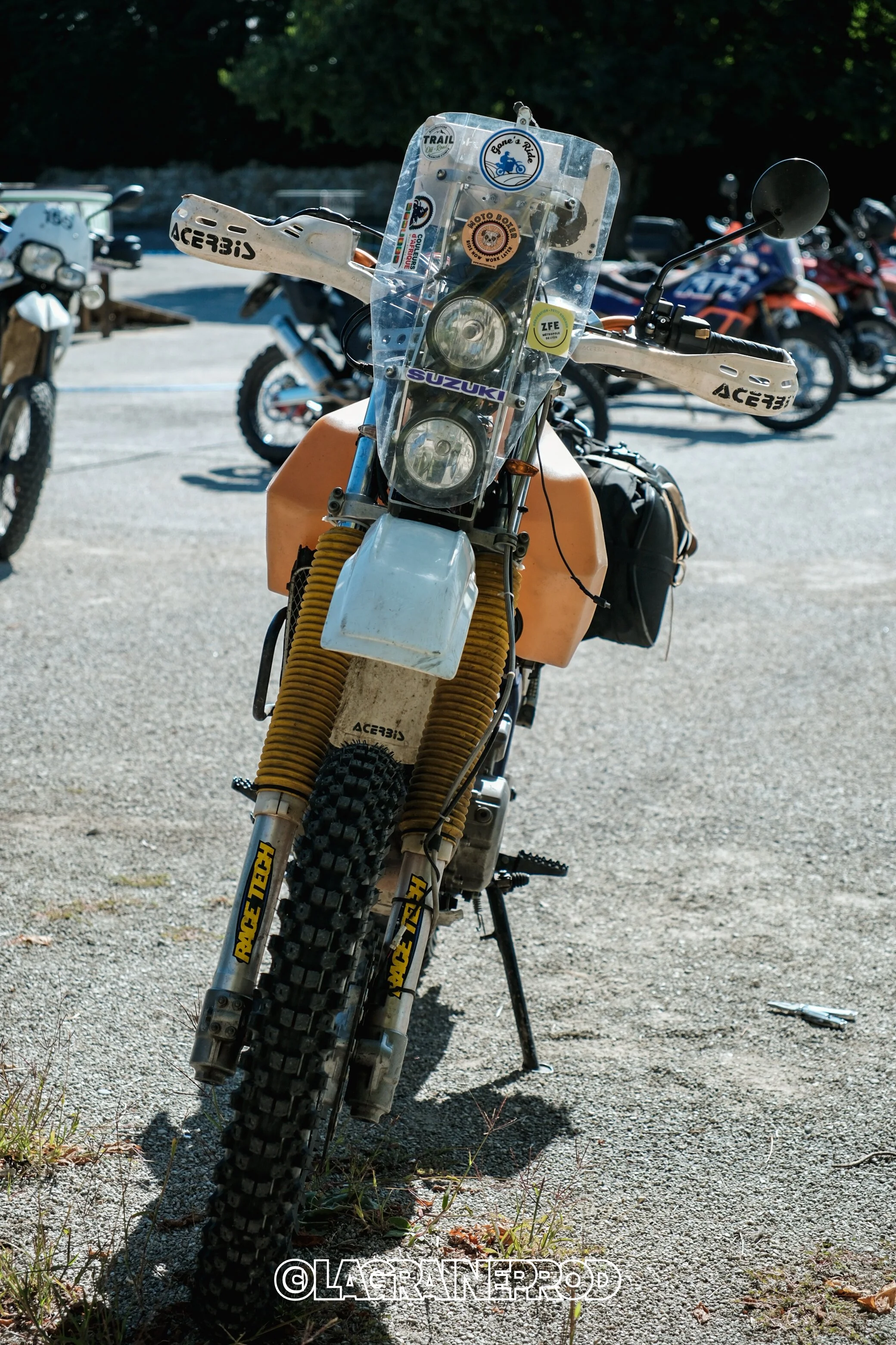 Une moto tout-terrain orange avec un pare-brise transparent, des pneus à crampons, et divers stickers, stationnée sur un terrain terreux avec d'autres motos en arrière-plan.