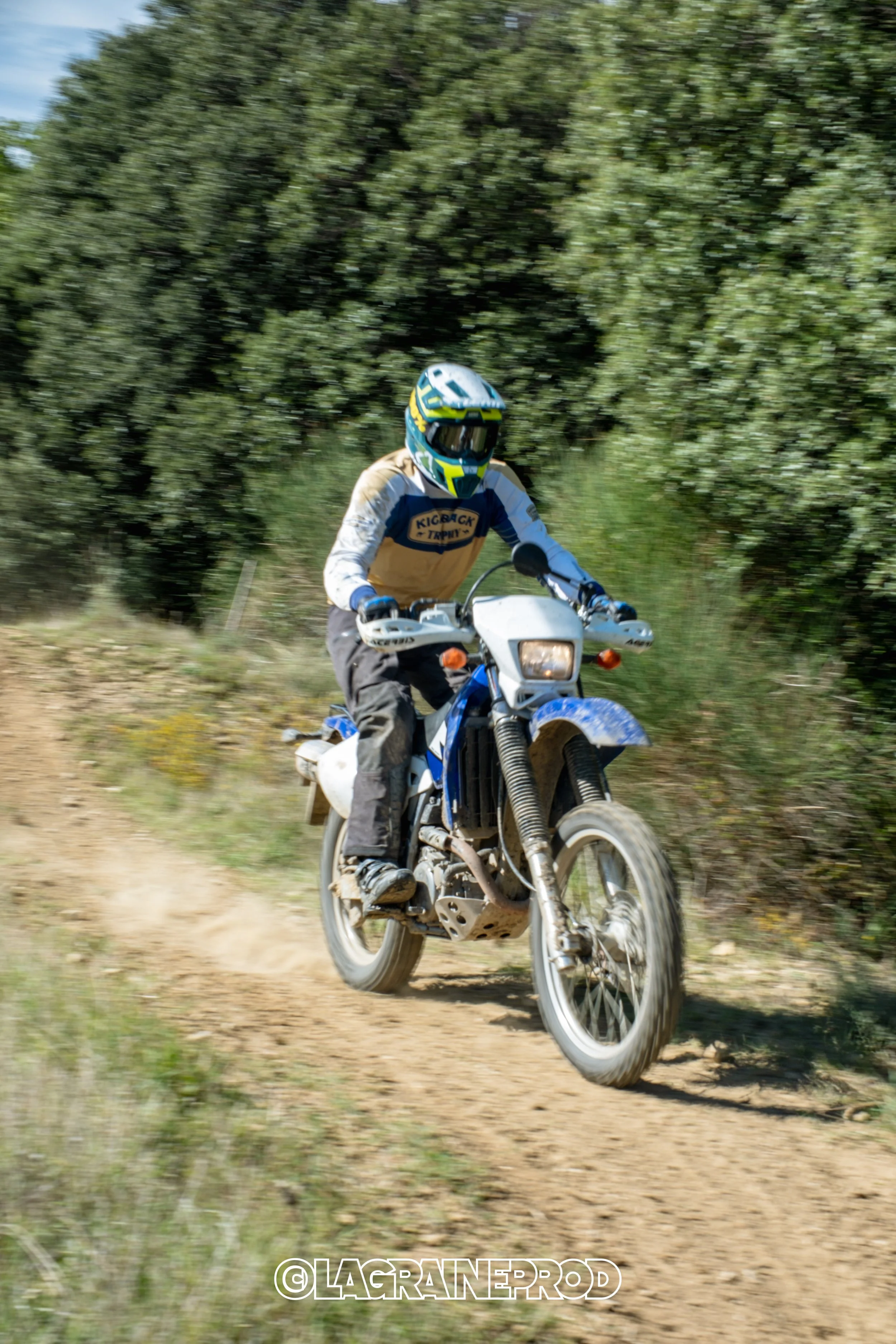 Un motard portant un casque bleu et des vêtements de protection, chevauchant une moto tout-terrain sur un sentier en dirt, avec des arbres verts en arrière-plan.