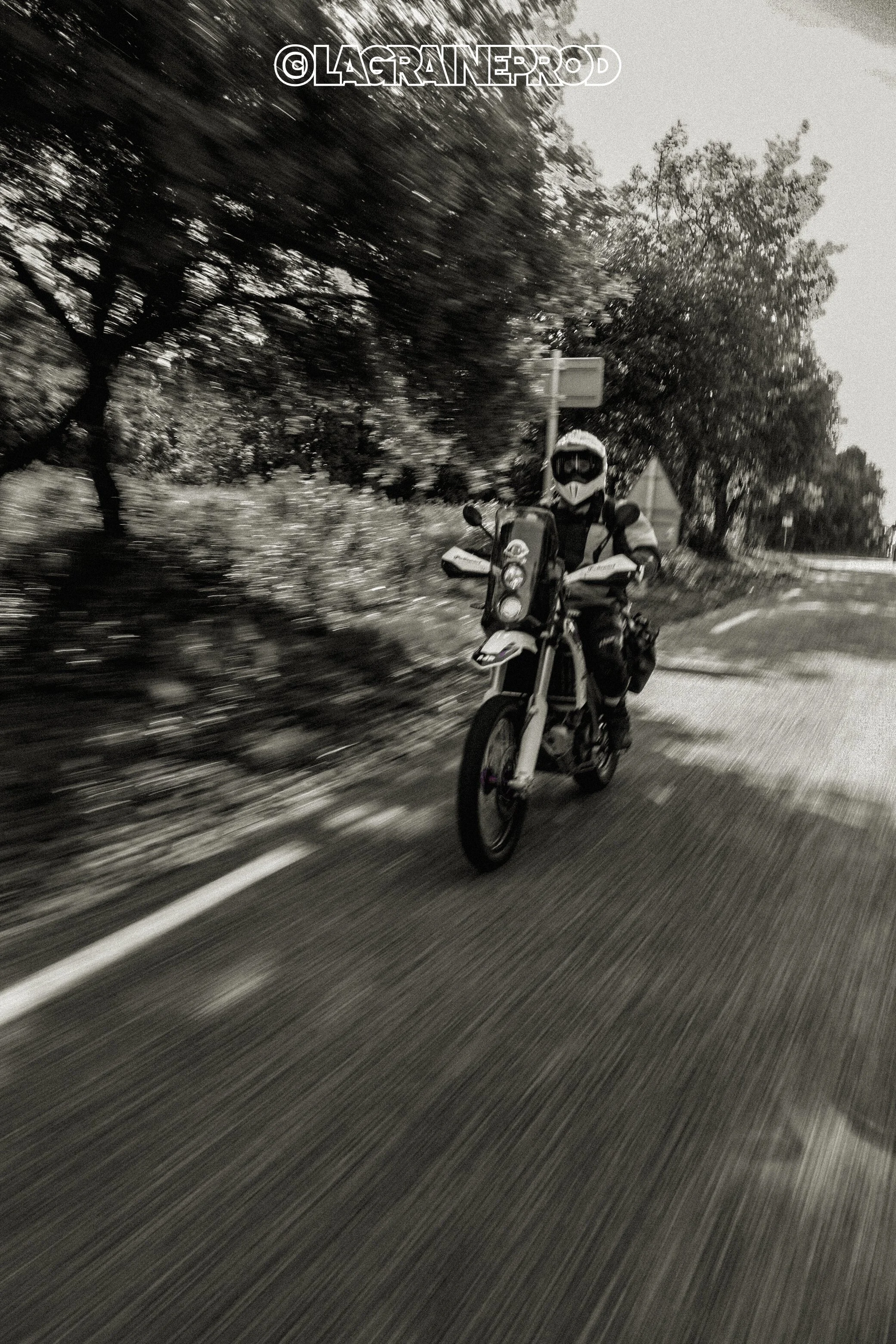 Motard avec casque et équipement de protection roulant sur une route bordée d'arbres, vue en noir et blanc