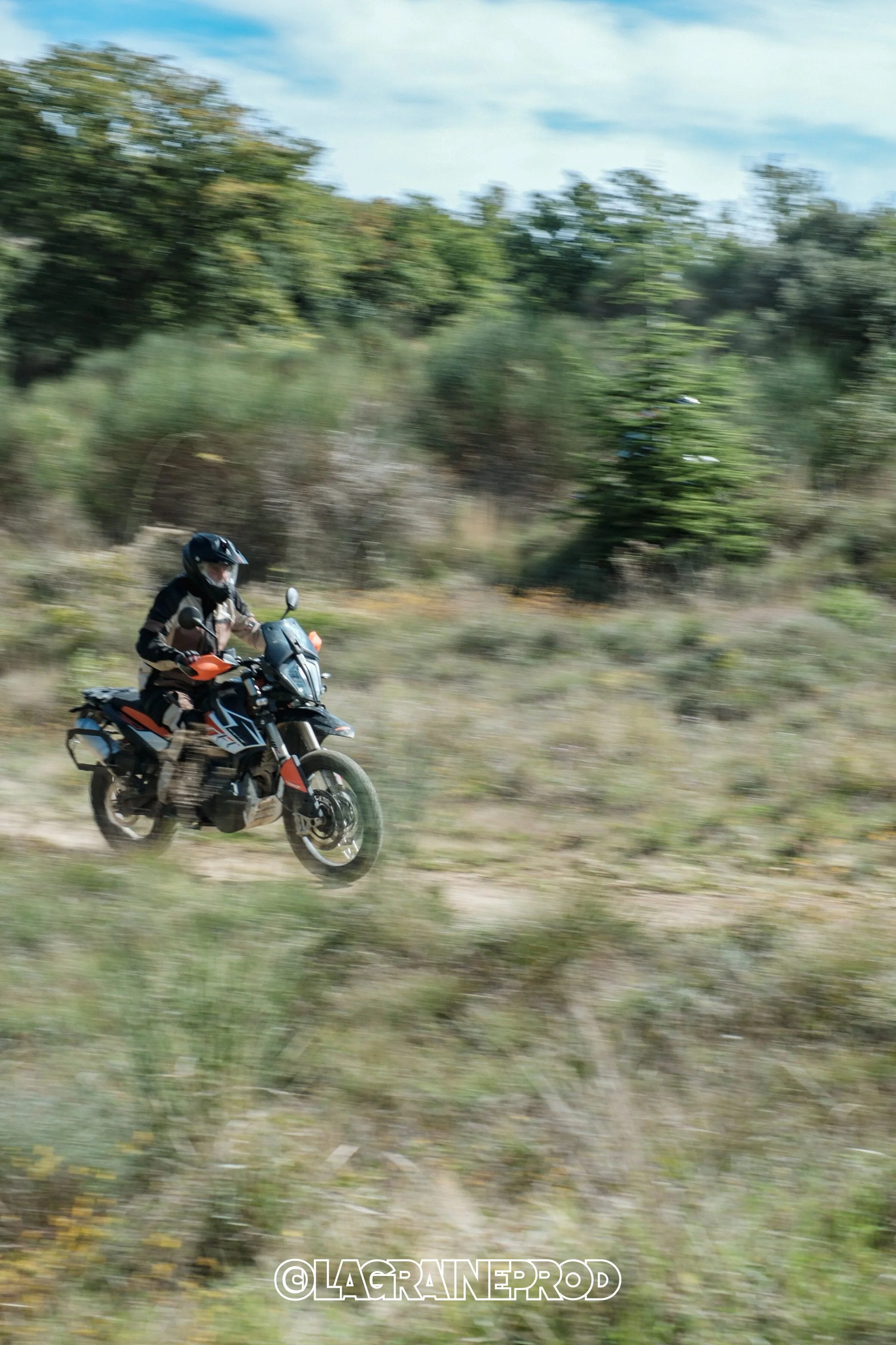 Un motard portant un casque noir et un équipement de protection, roulant à moto tout-terrain sur un chemin bordé d'arbres verts, avec un ciel partiellement nuageux.