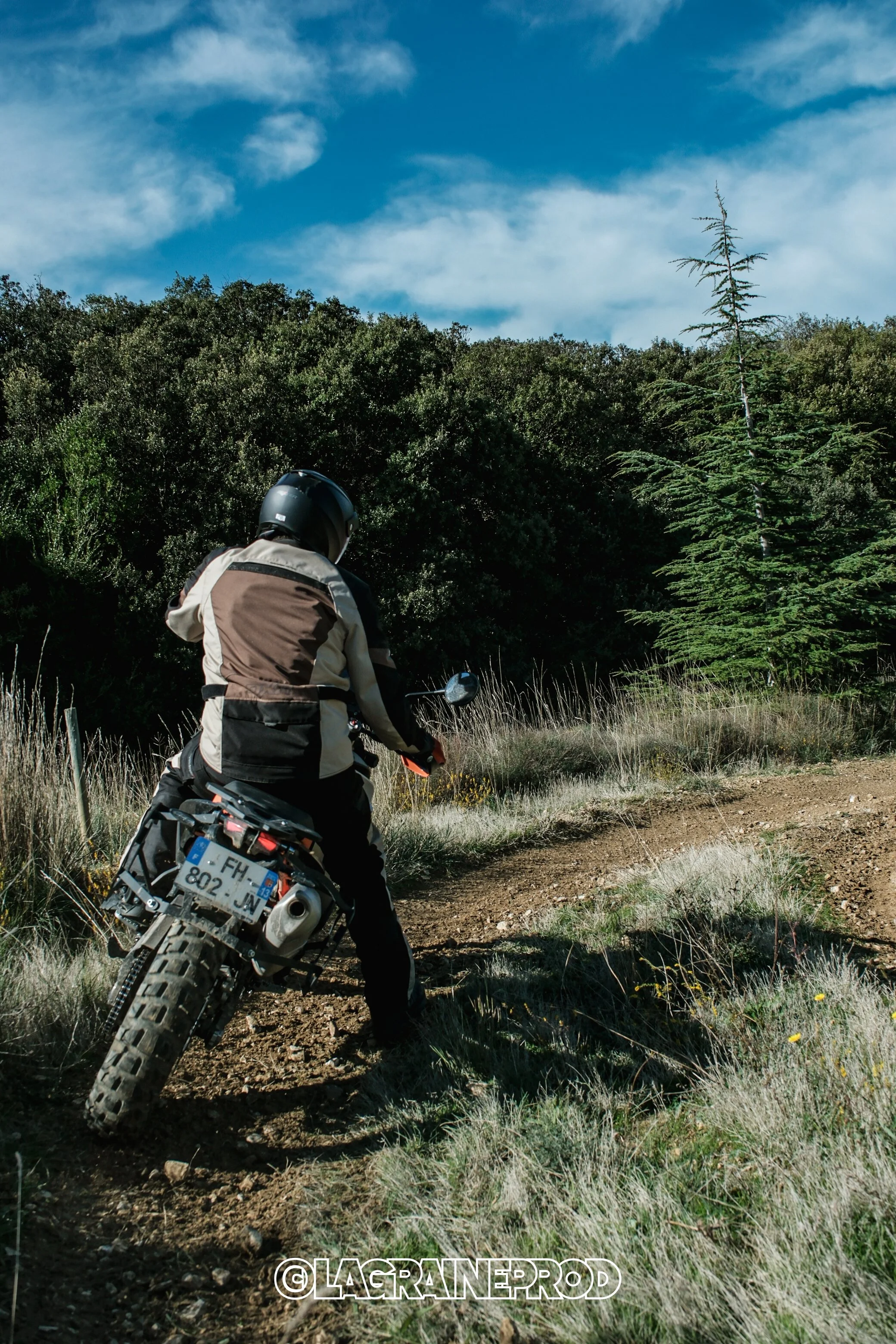 Un motard portant un casque et un équipement de moto tout-terrain, en train de rouler sur un sentier en pleine nature, avec des arbres et un ciel bleu en arrière-plan.