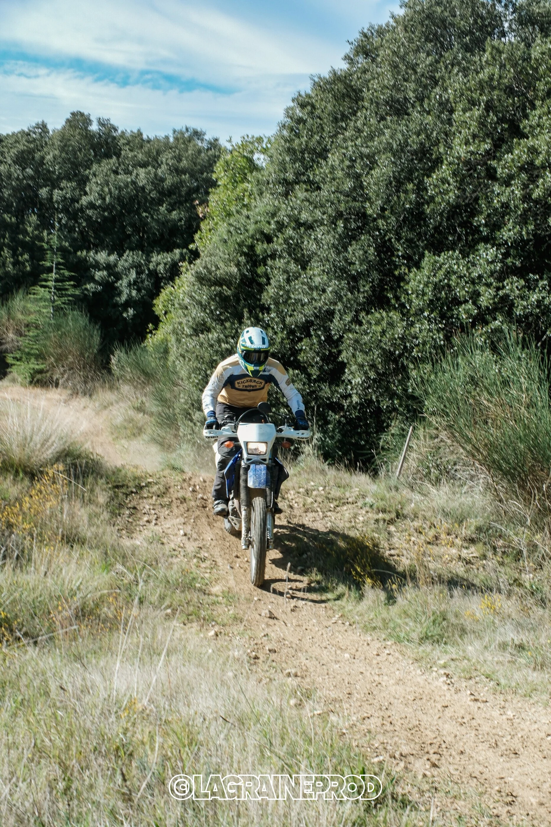 Un motocycliste portant un casque et des vêtements de protection, roulant sur un sentier de terre dans une zone verdoyante avec des arbres et du ciel bleu.