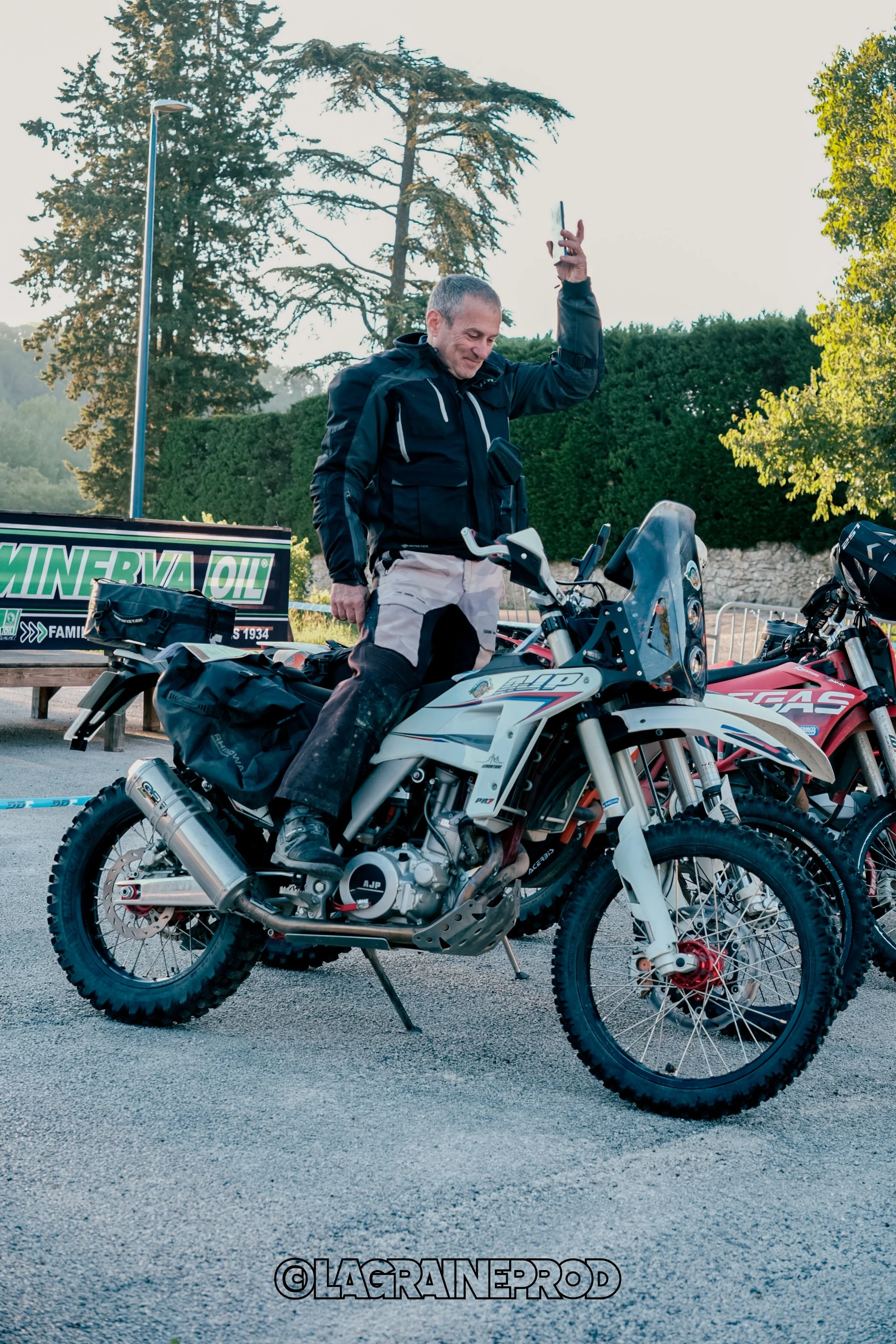 Homme souriant en tenue de moto, assis sur une moto d'aventure, levant la main en saluant, avec deux motos visibles et un arbre en arrière-plan.