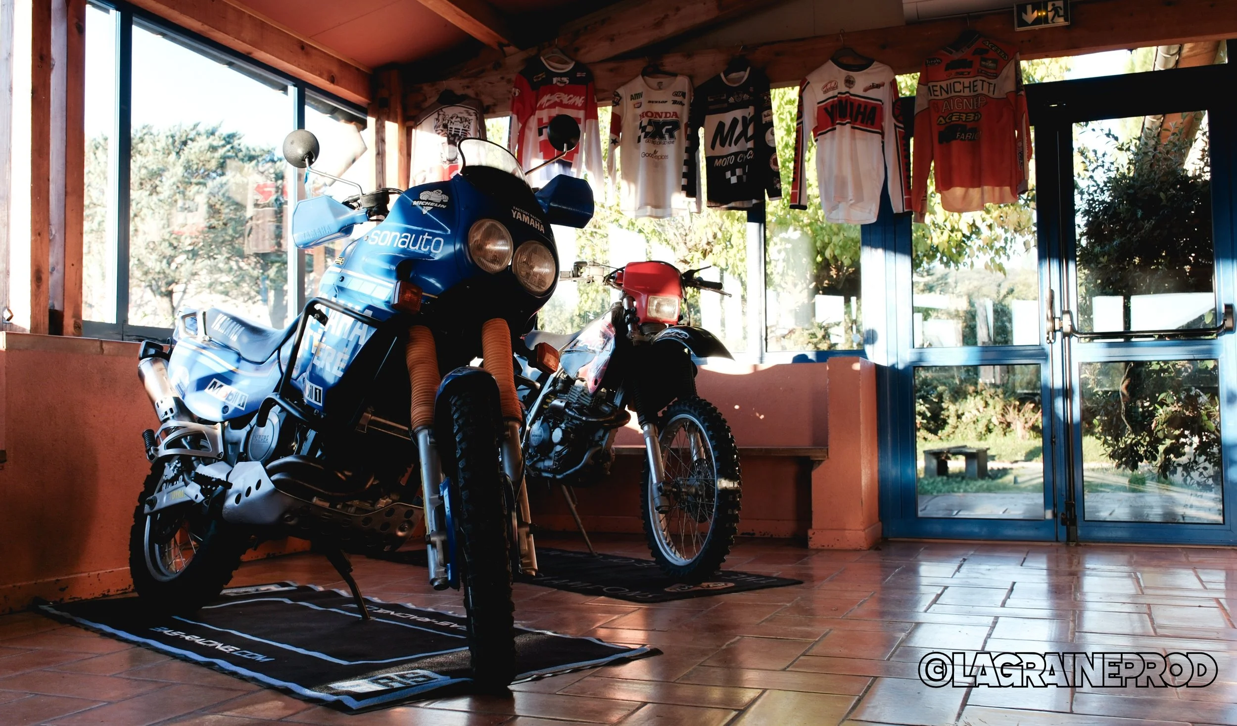 Deux motocyclettes anciennes, une bleue et une rouge, exposées à l'intérieur d'une pièce avec de grandes fenêtres et des vêtements de motocross suspendus au plafond, vue de l'intérieur avec porte en verre menant à l'extérieur.
