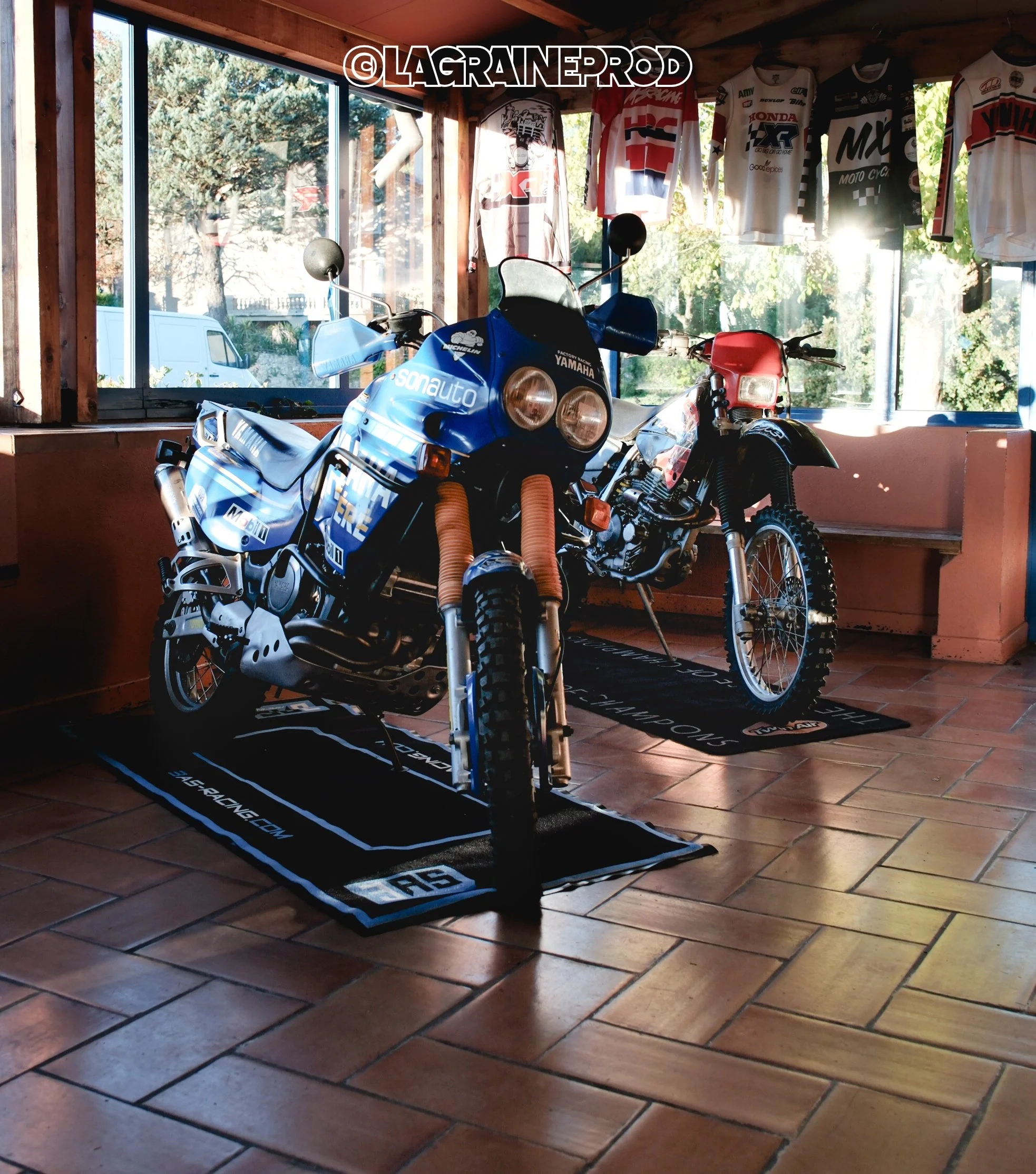Deux motos de motocross dans une pièce, avec des vestes et t-shirts suspendus au mur en arrière-plan, et une grande fenêtre laissant entrer la lumière naturelle.