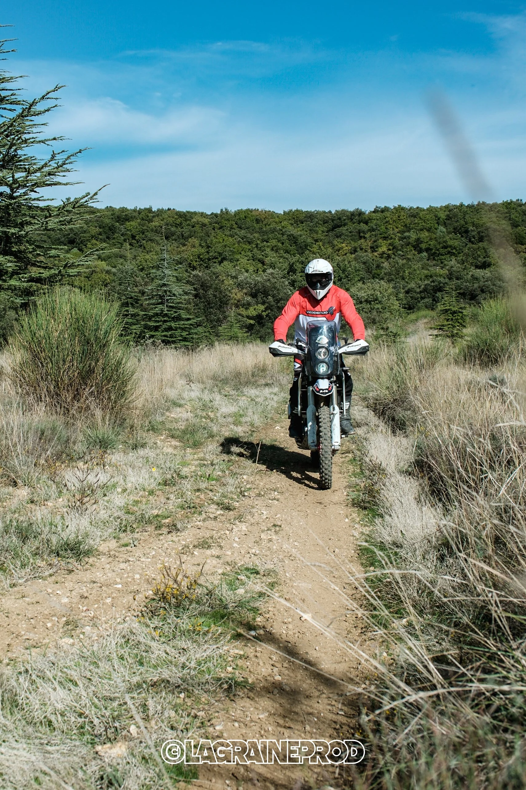 Personne conduisant une moto tout-terrain sur un sentier de terre dans un paysage naturel avec des arbres et un ciel bleu.