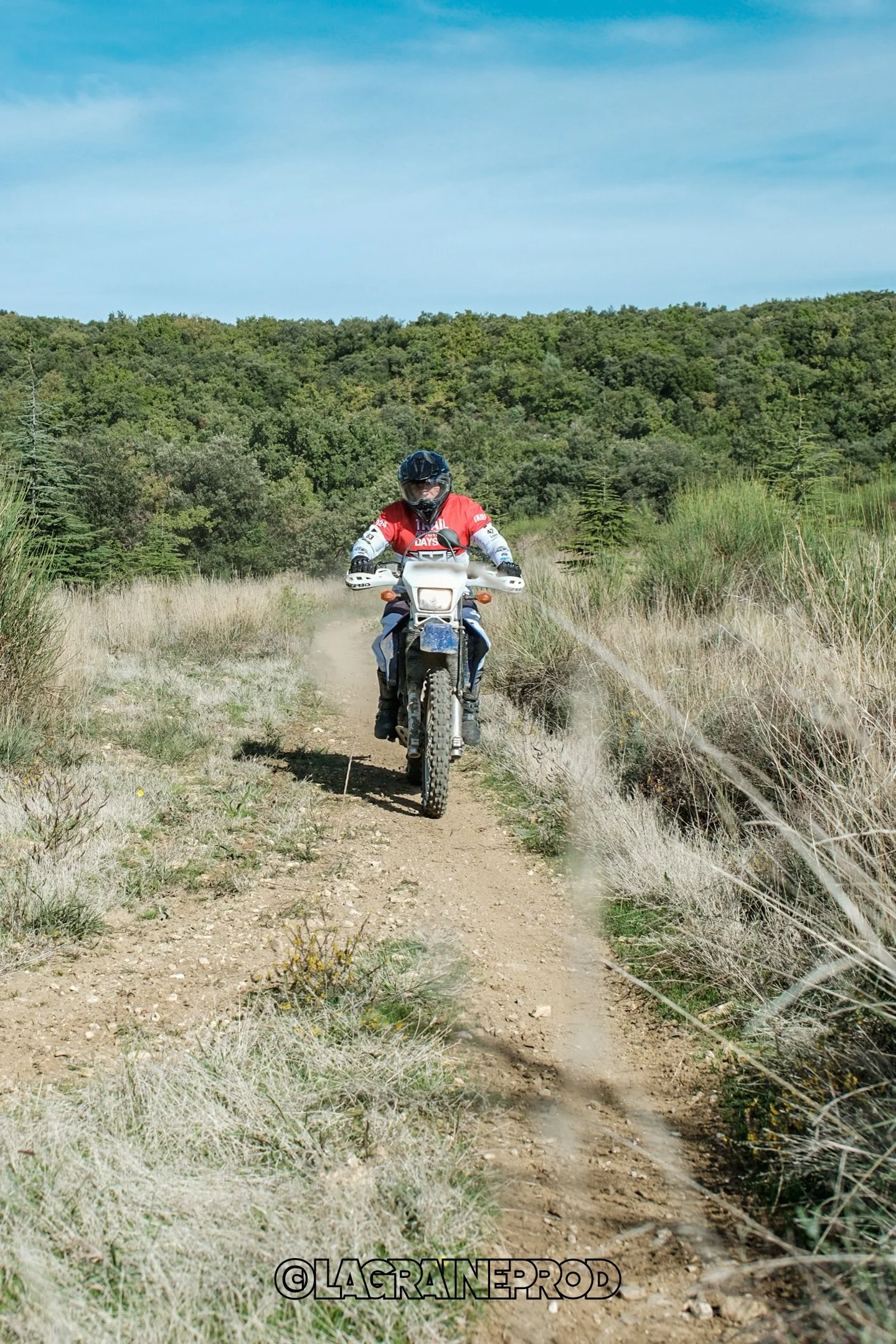Un motard portant un casque noir et une veste rouge, rouge et blanche, roulant sur une piste de terre en pleine nature, entourée d'herbes et d'arbres, sous un ciel bleu.