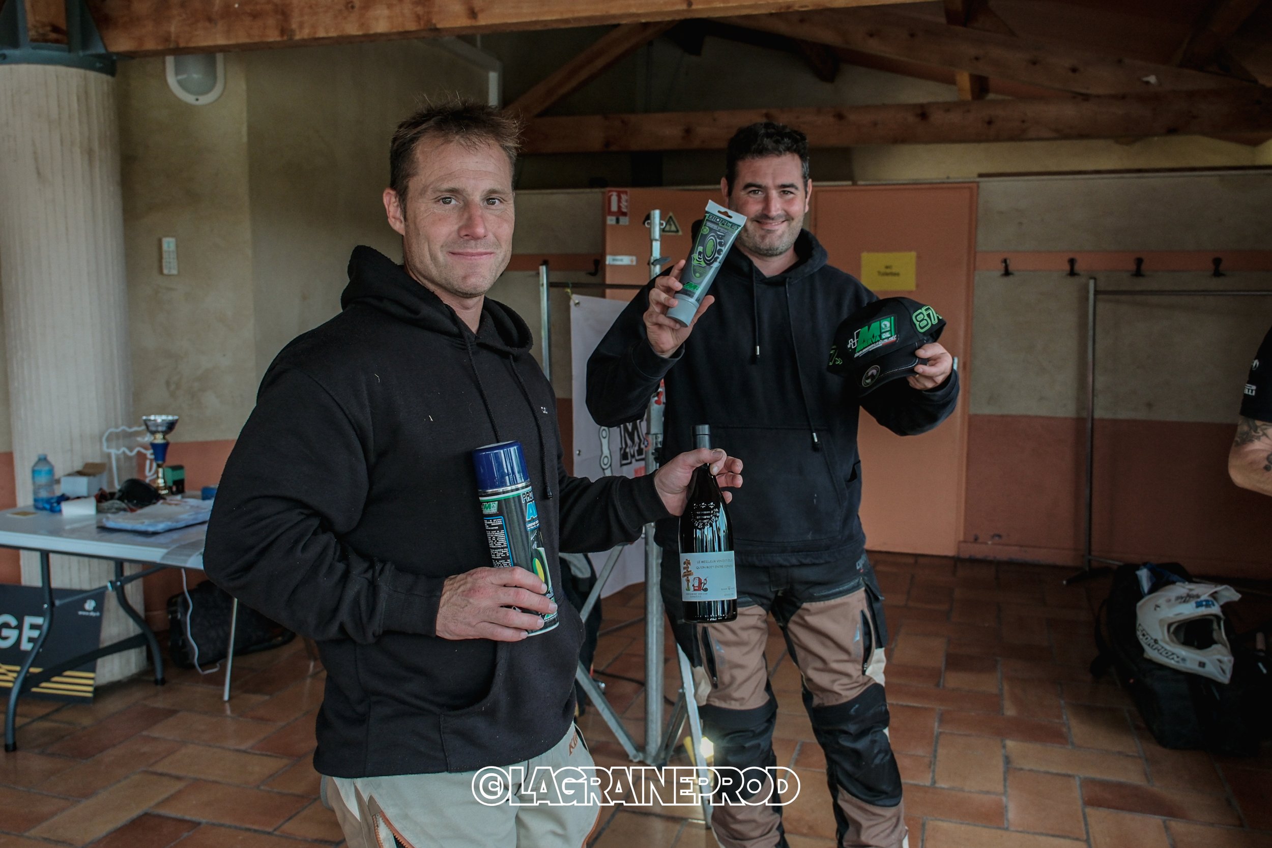 Deux hommes souriants en tenue de moto, tenant des bouteilles de vin et des produits pour la moto, dans une pièce avec un sol en carreaux et un plafond en bois.