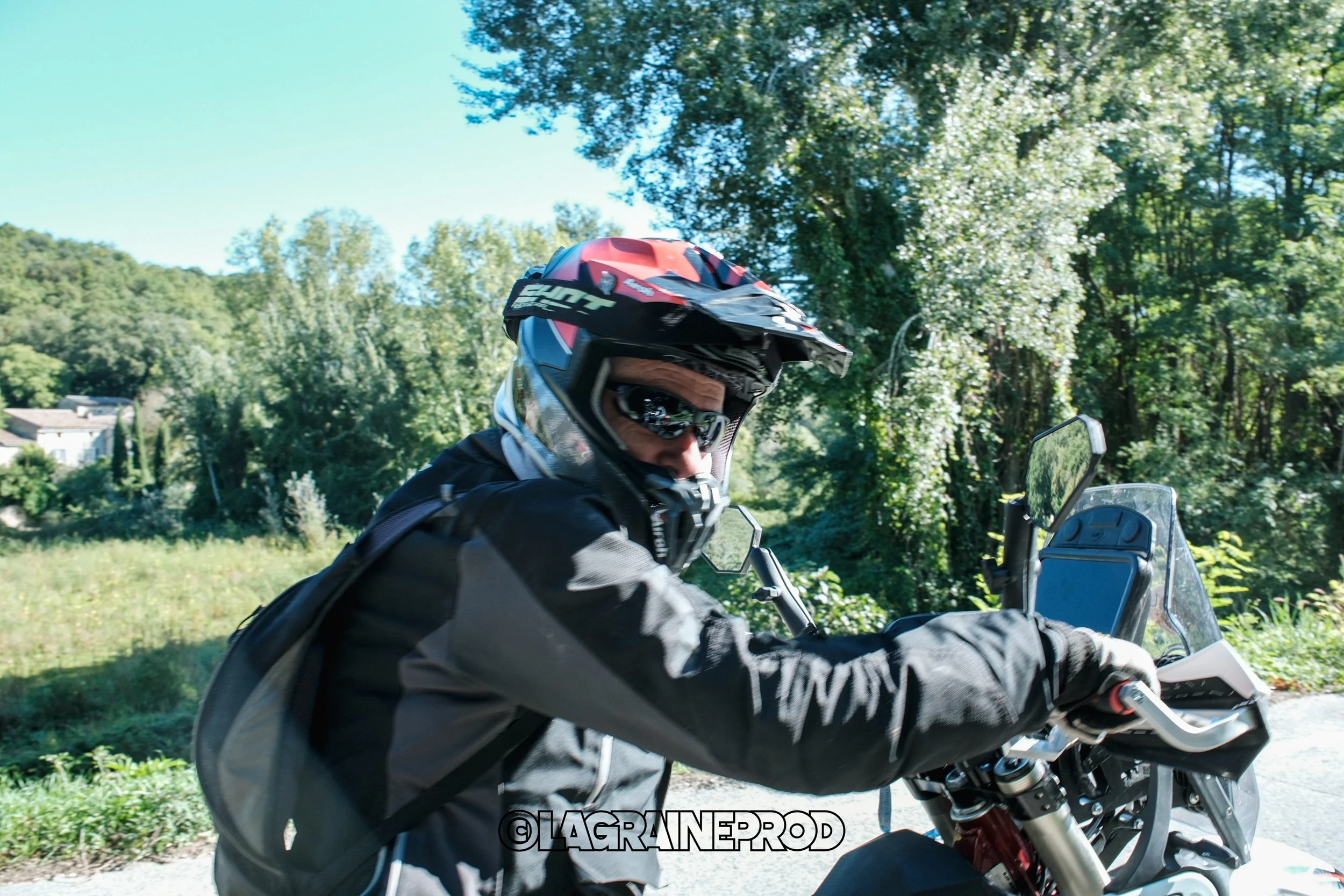 Un homme portant un casque de moto et des vêtements de protection, assis sur une moto dans un environnement naturel verdoyant.