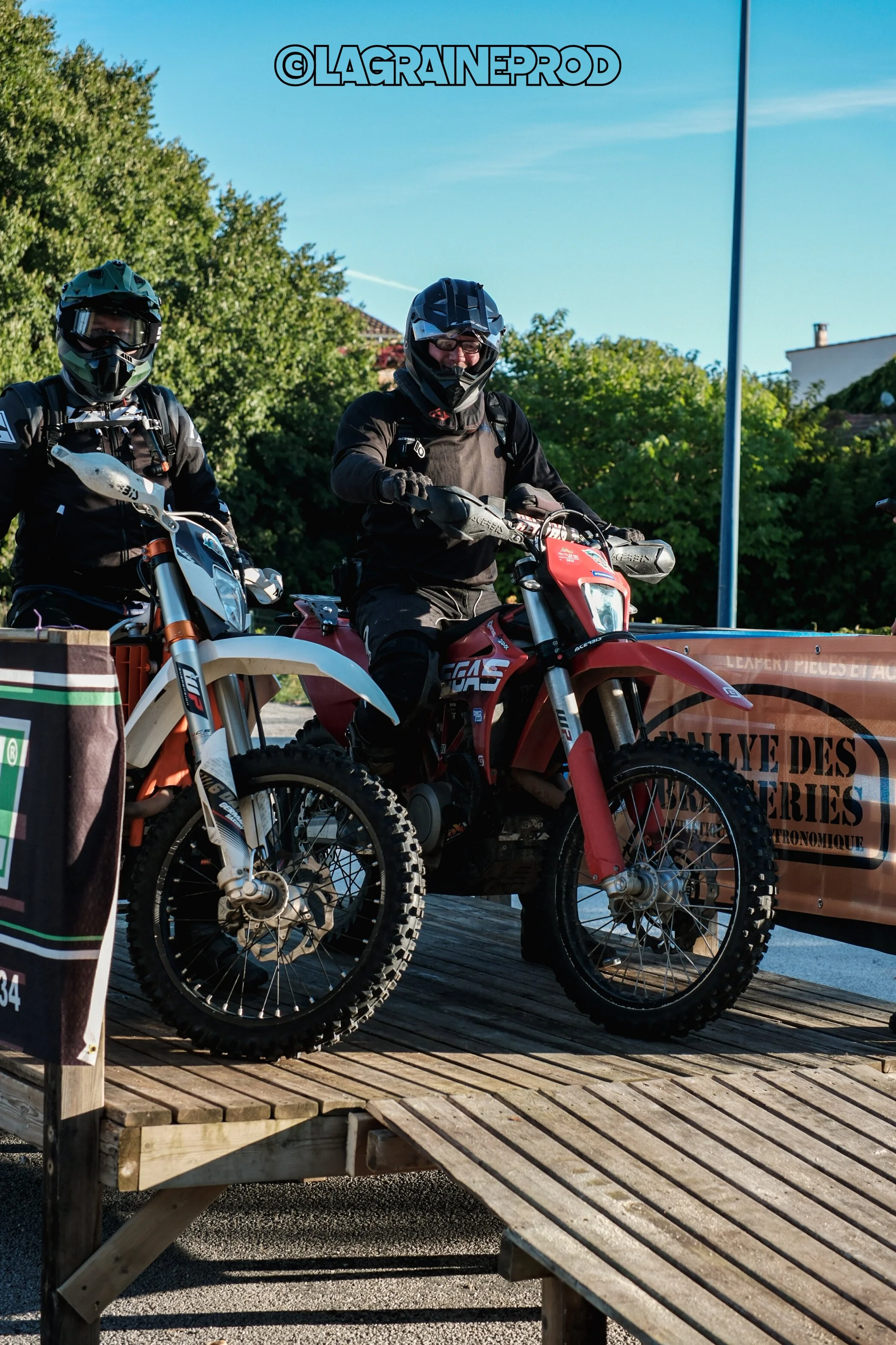 Deux motocyclistes portant des casques et des équipements de protection, côte à côte avec des motos tout-terrain, sur une plateforme en bois lors d'une compétition ou d'une sortie en plein air.