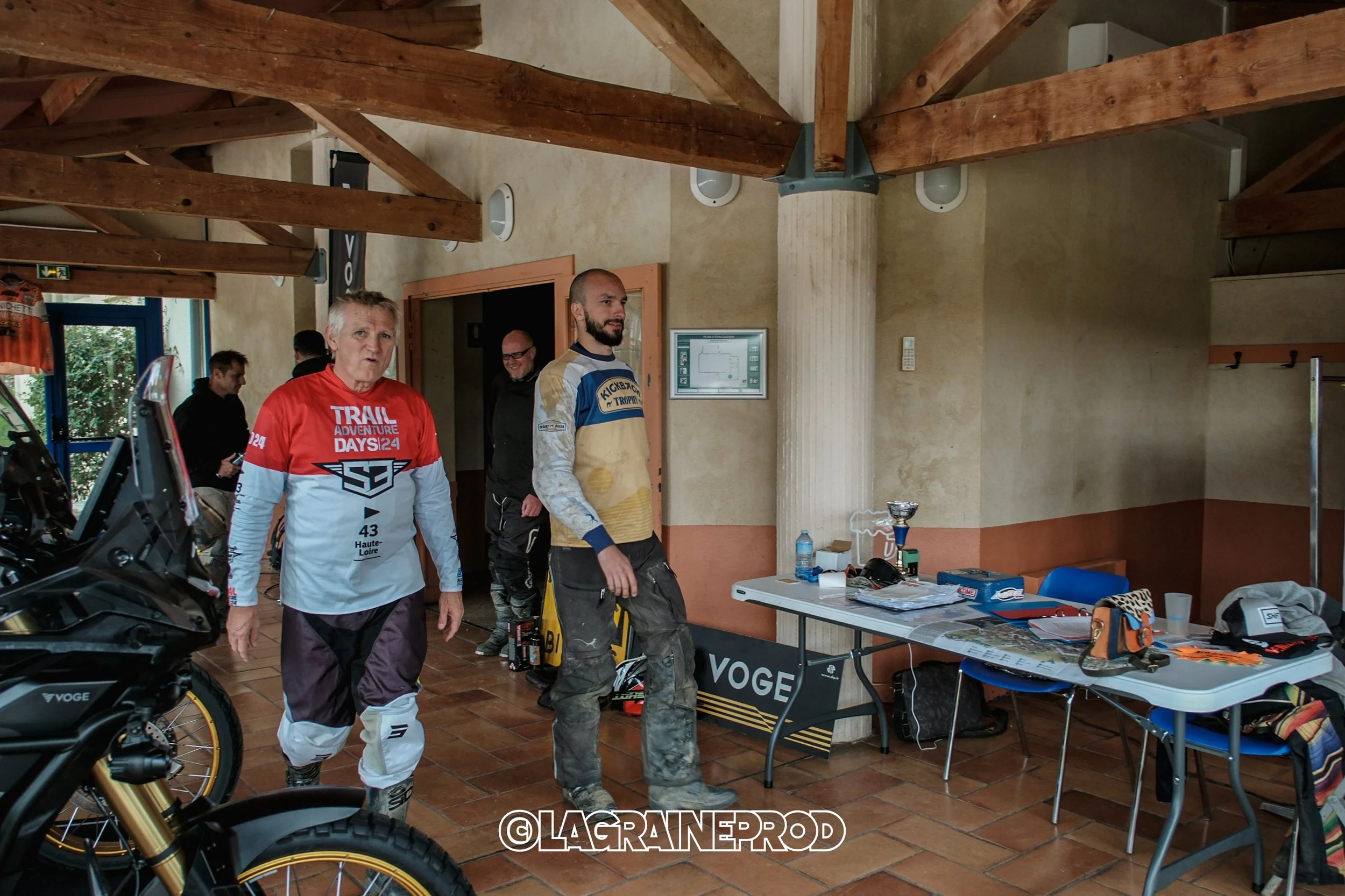 Intérieur d'une salle avec plusieurs hommes, un d'entre eux porte un maillot de motocross rouge, blanc et noir. La pièce a des murs beige et un plafond en bois avec des poutres. Une table blanche est couverte de divers objets, y compris une bouteille