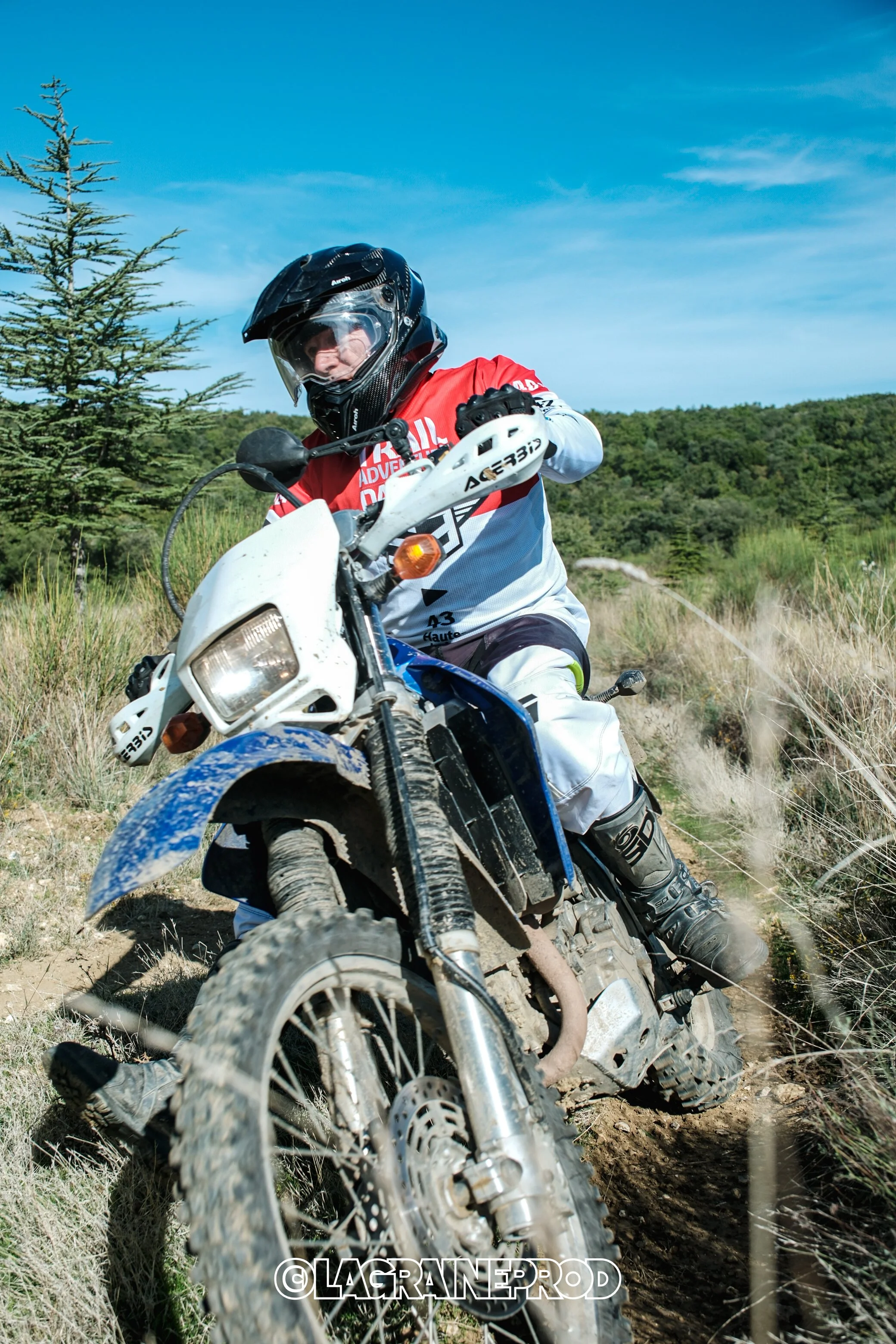 Un pilote de moto tout-terrain portant un casque intégral, une chemise rouge et blanche, en train de naviguer sur un sentier rocheux en plein air, avec un ciel bleu et des arbres en arrière-plan.
