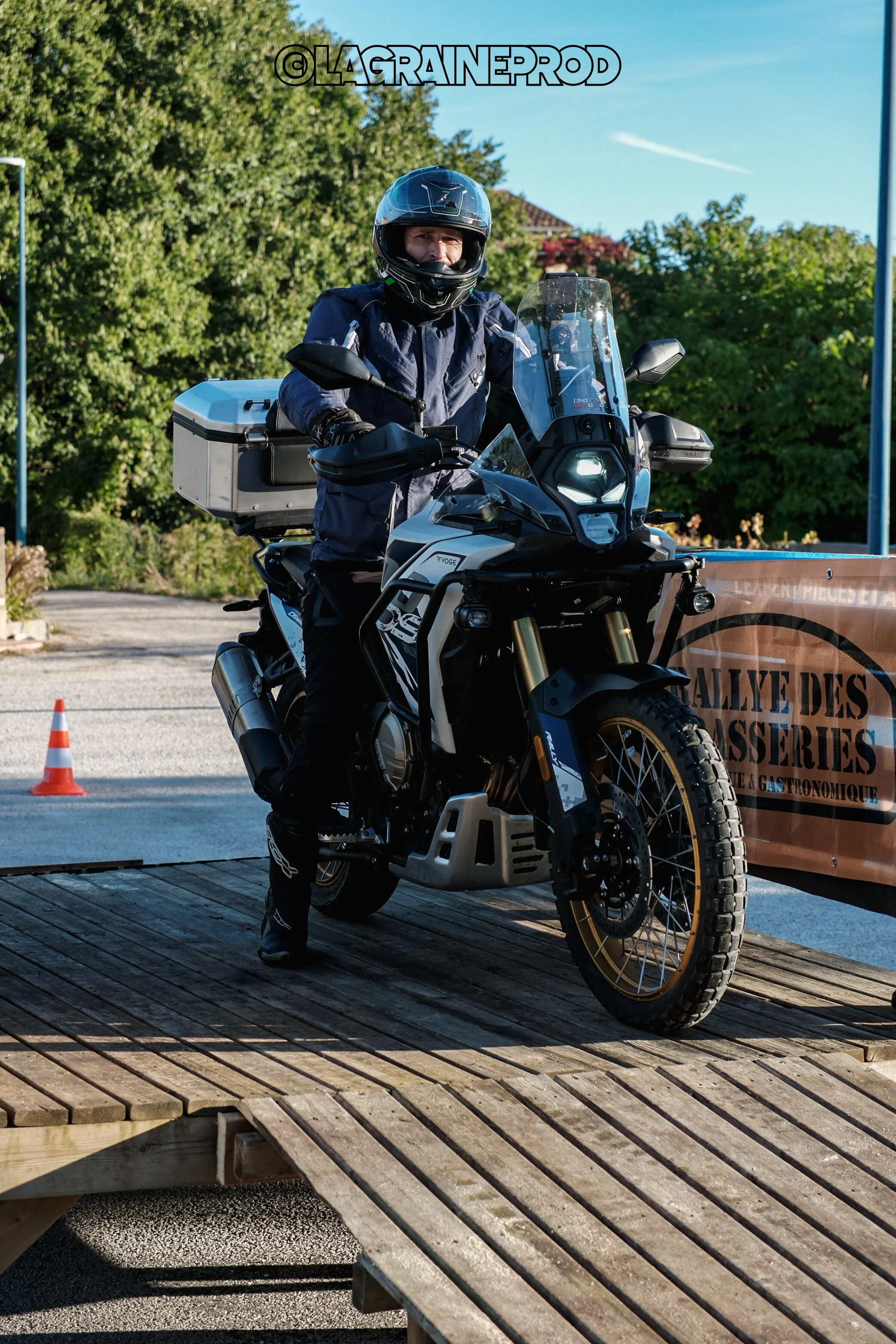 Motard en tenue de protection assis sur sa moto tout-terrain, portant un casque intégral, avec un panneau annonçant un rallye.