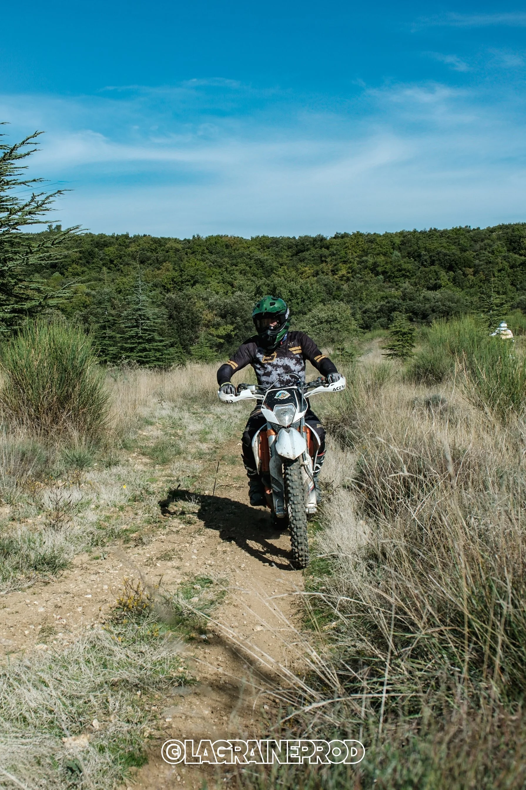 Un motard portant un casque et une tenue de protection, roulant sur une moto tout-terrain sur un chemin de terre dans une nature verdoyante avec des arbres et un ciel bleu.