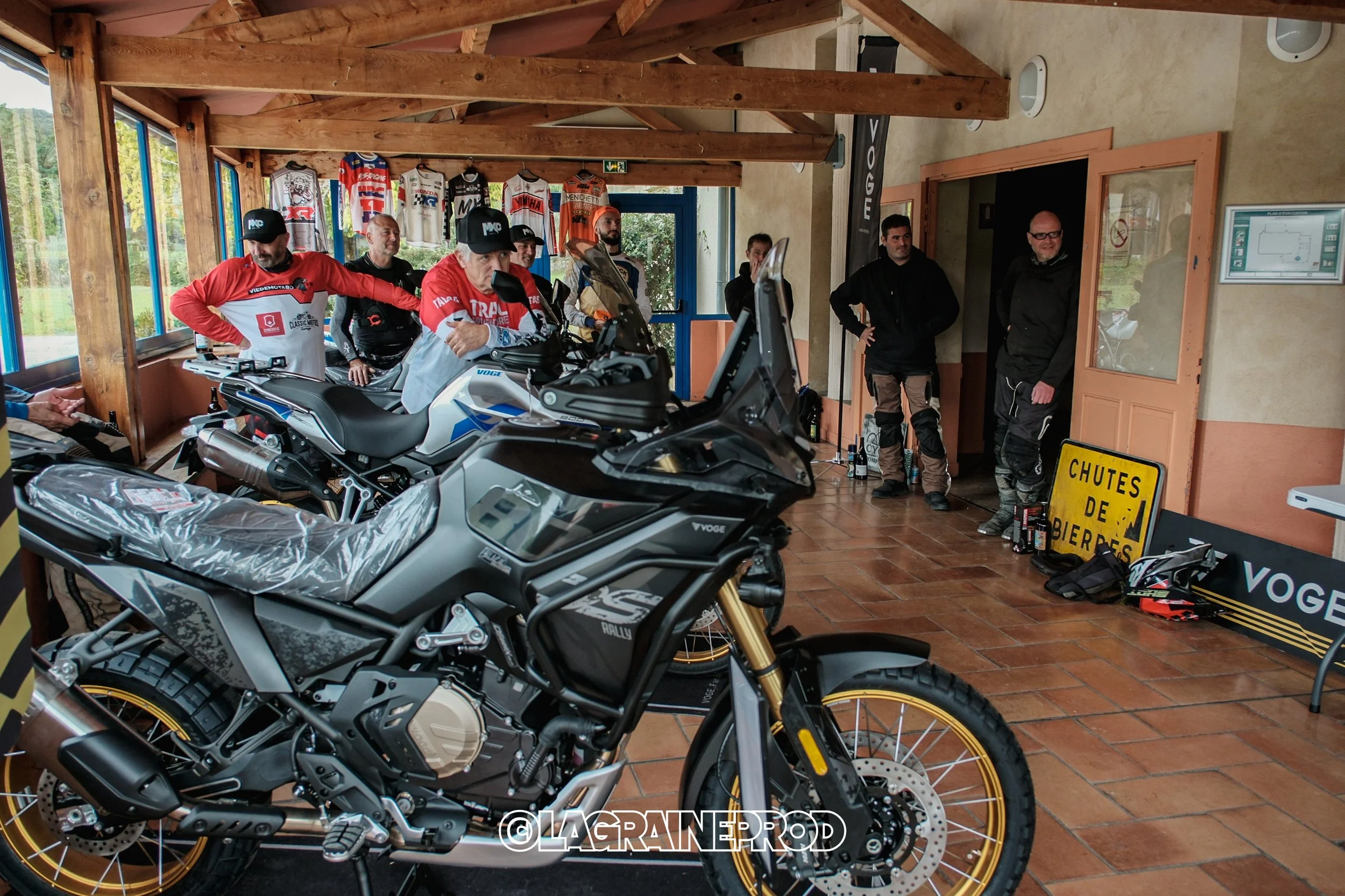 Un groupe de motards dans une pièce, avec plusieurs motos Voge en avant-plan, des casques, des bouteilles, et un panneau jaune indiquant 'Chutes de pierres'.