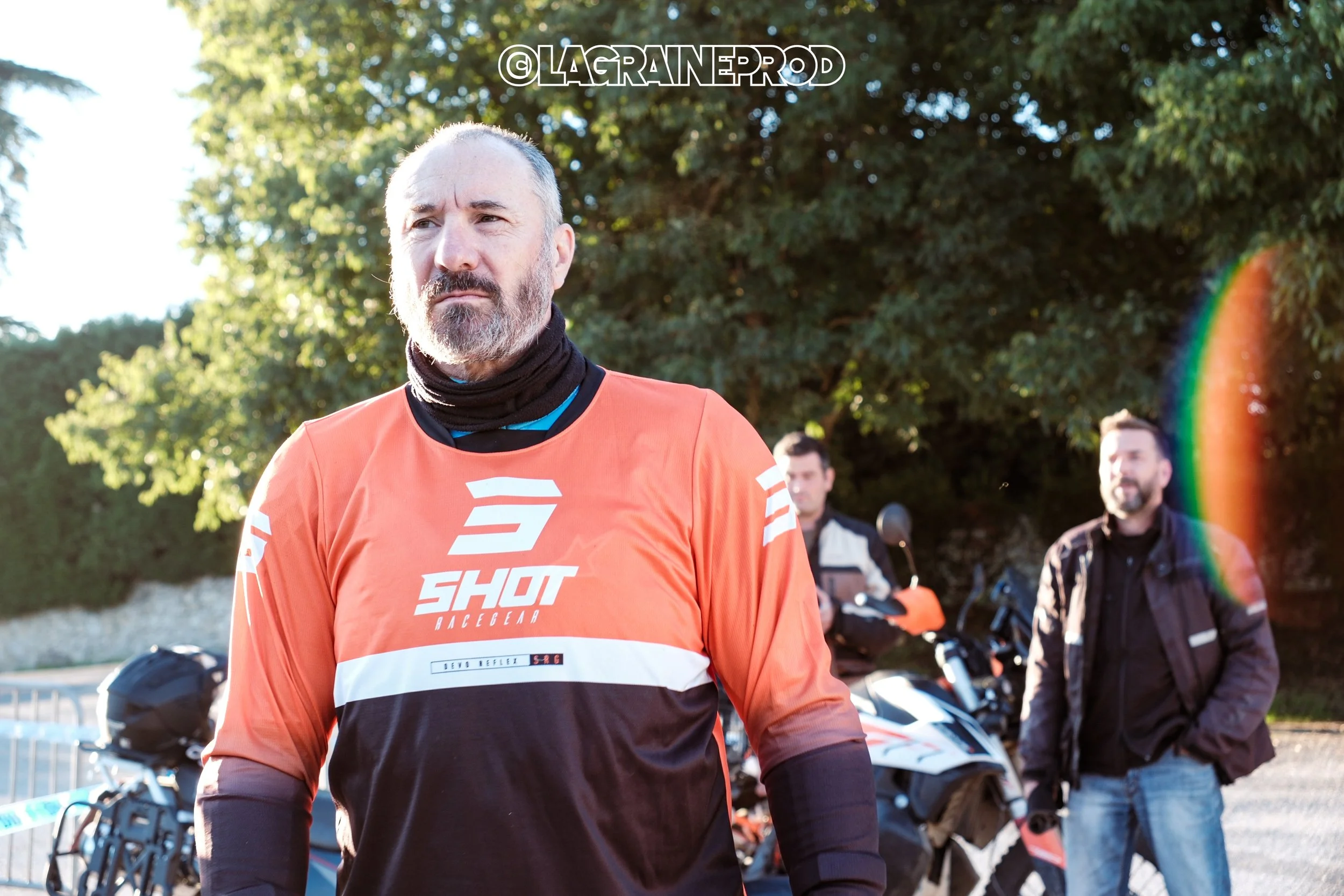 Groupe de trois personnes à l'extérieur avec des motos, avec un homme en premier plan portant un maillot orange avec le logo 'SHOT'.