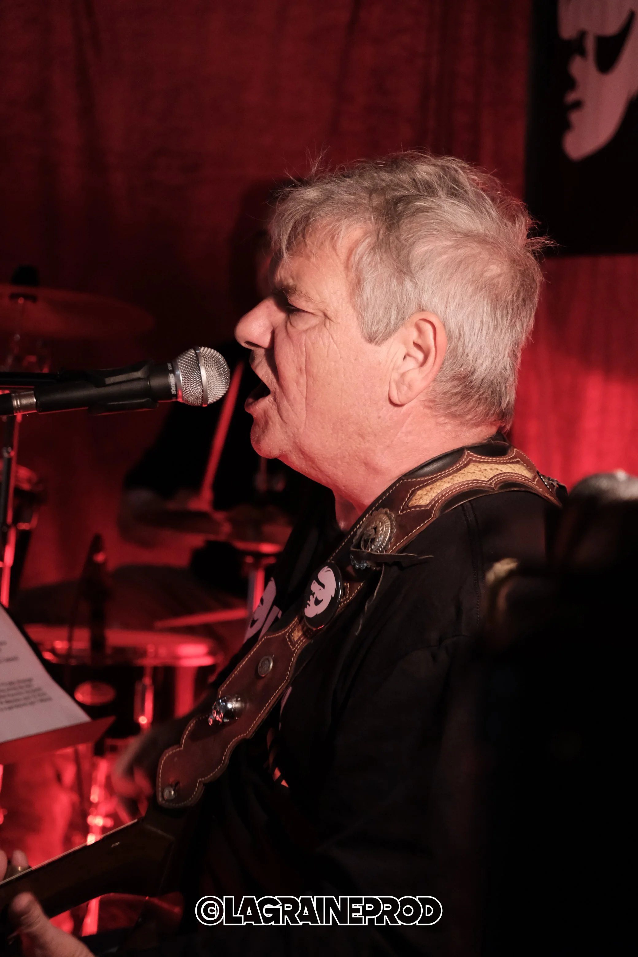 Un homme chante en jouant de la guitare dans un lieu avec un éclairage rouge, un micro devant lui, et un décor noir et rouge en arrière-plan.