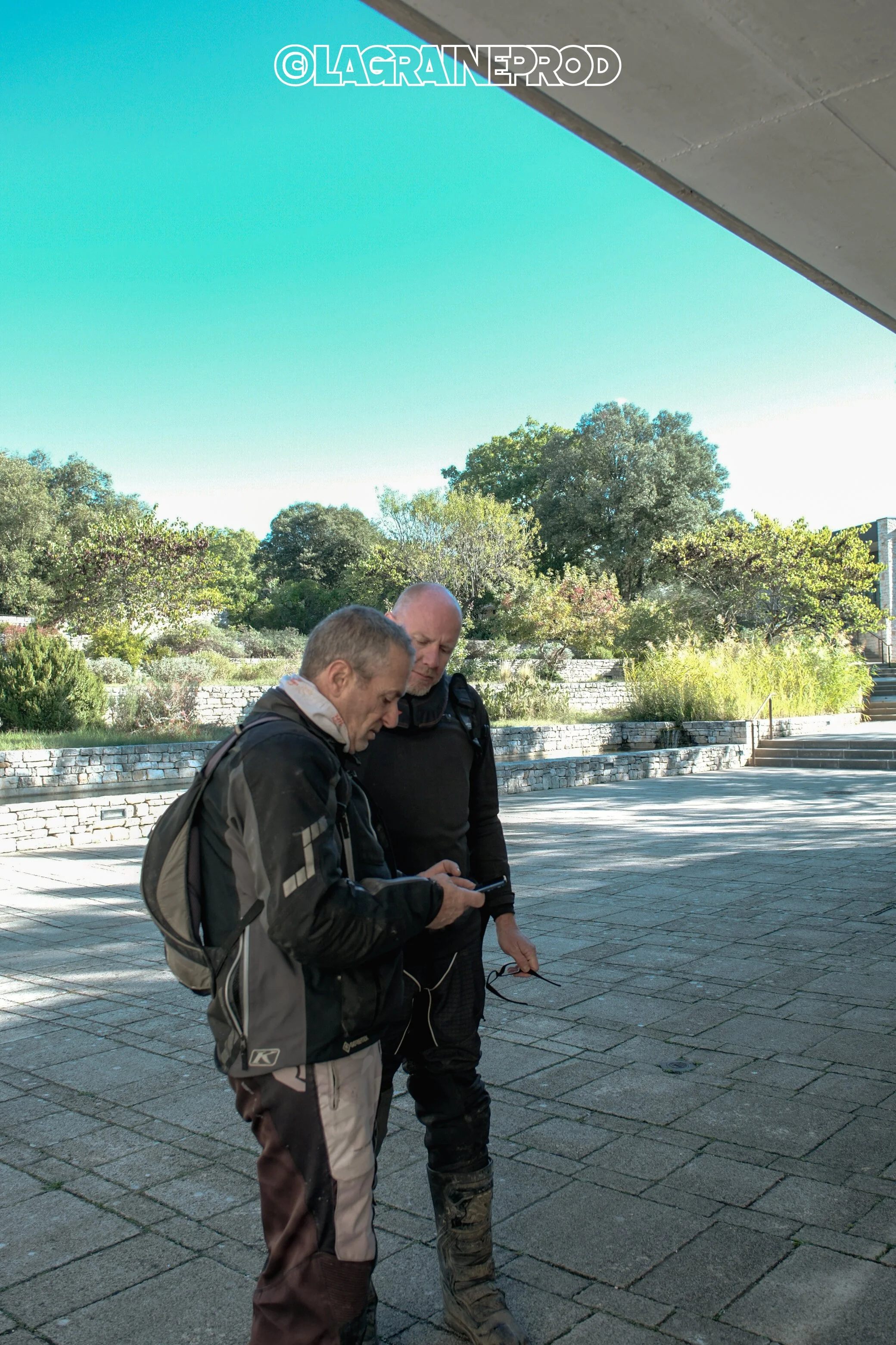 Deux hommes regardent leur téléphone portable en plein air sous un ciel clair, avec des arbres et une zone pavée en arrière-plan.
