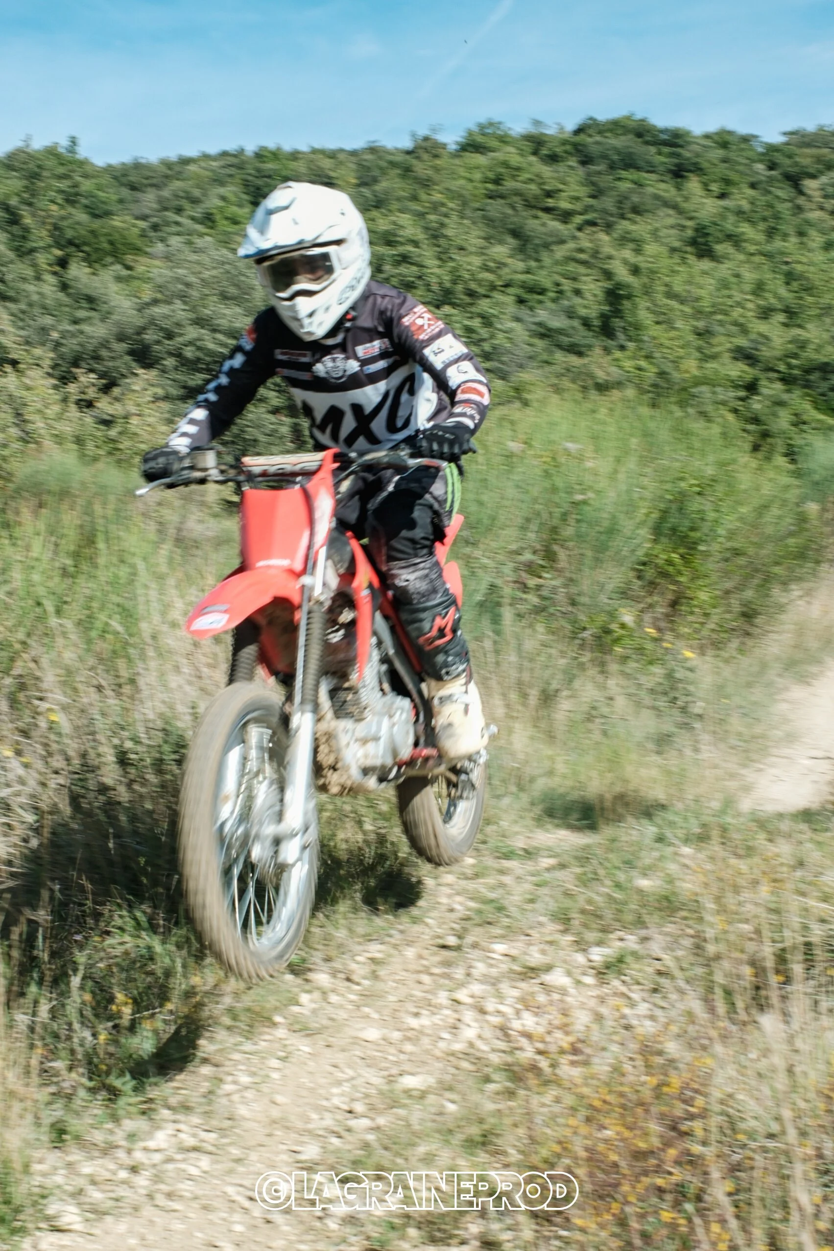 Coureur de motocross sur une piste en plein air, portant un casque blanc, une tenue noire et un équipement de protection, sur une moto rouge dans un décor naturel avec des arbustes et un ciel bleu.