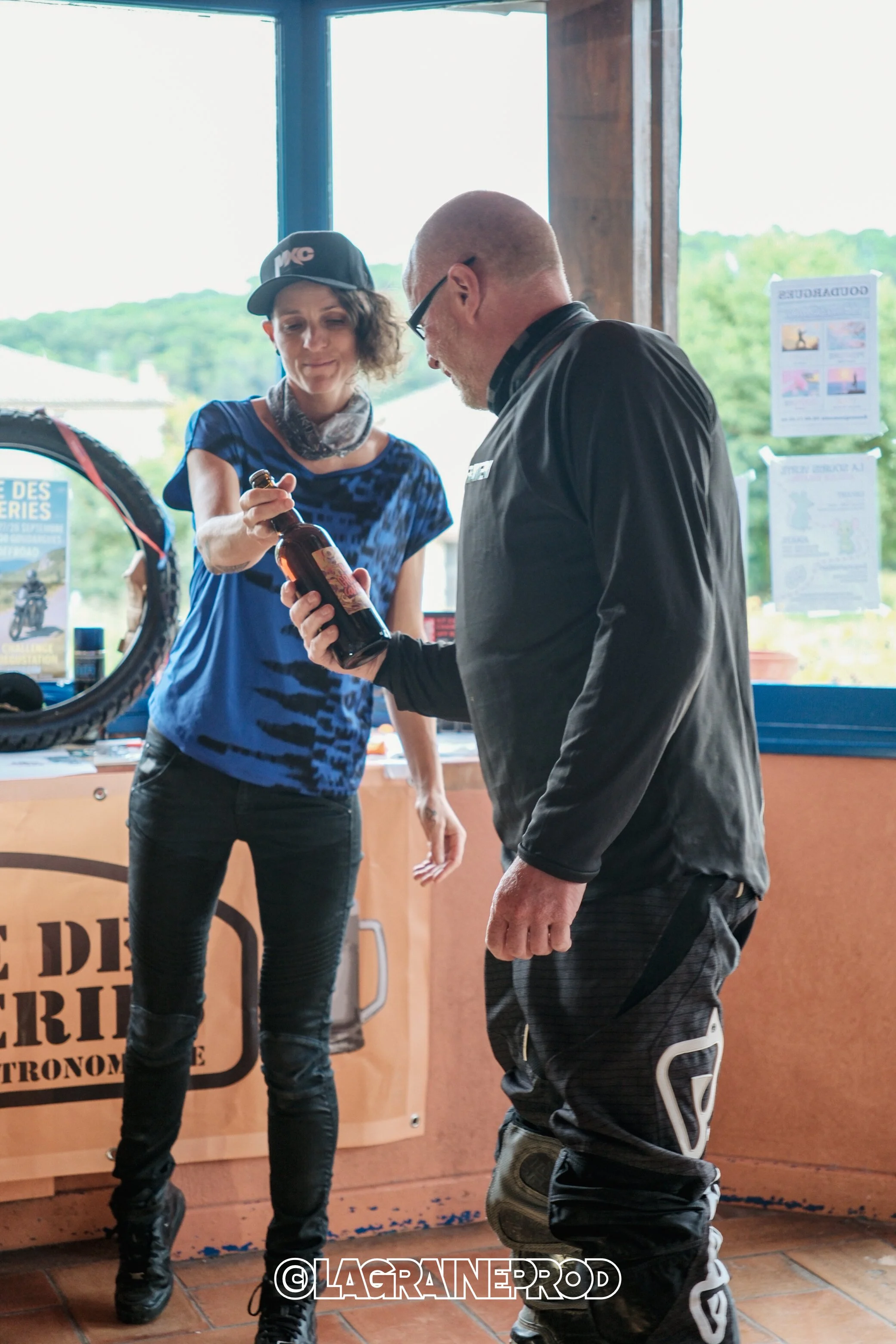 Une femme tend une bouteille à un homme dans un magasin ou une boutique. La scène se déroule près d'une fenêtre avec un paysage vert en arrière-plan.