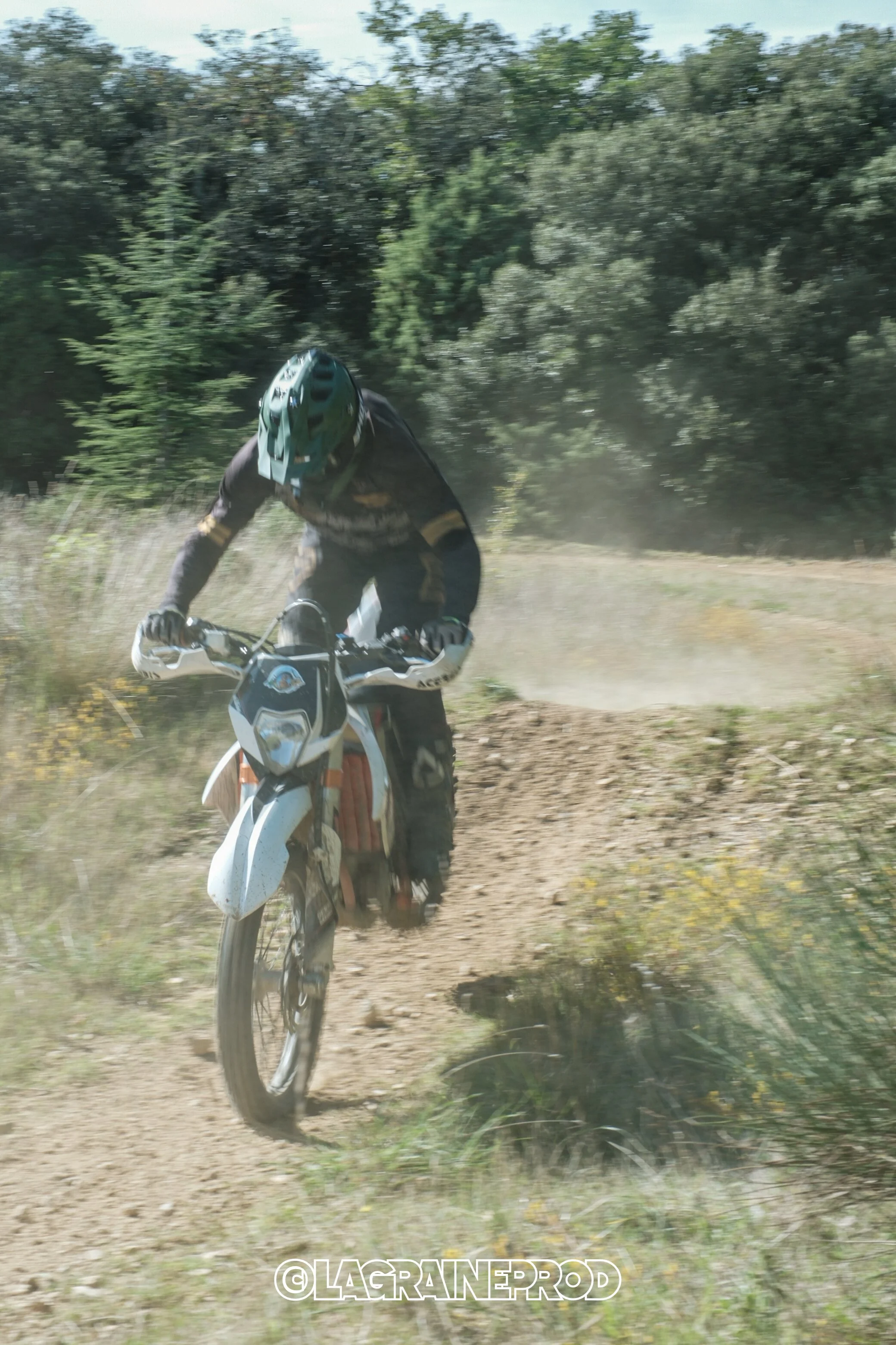 Un motocycliste portant un casque et un costume de protection, roulant sur un sentier poussiéreux dans une zone boisée.